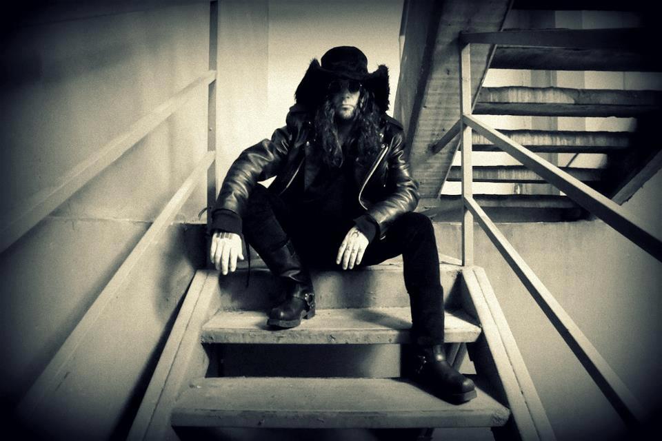 Imagen en blanco y negro del expresentador de televisión Luis Lauretto vestido con chamarra de cuero negra, pantalón negro y sombrero vaquero negro. Está sentado en unas gradas.