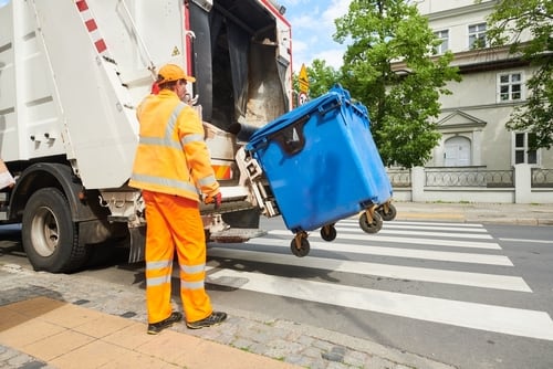 Trabajador municipal en uniforme naranja operando un camión de basura mientras recoge un contenedor azul en una calle urbana.