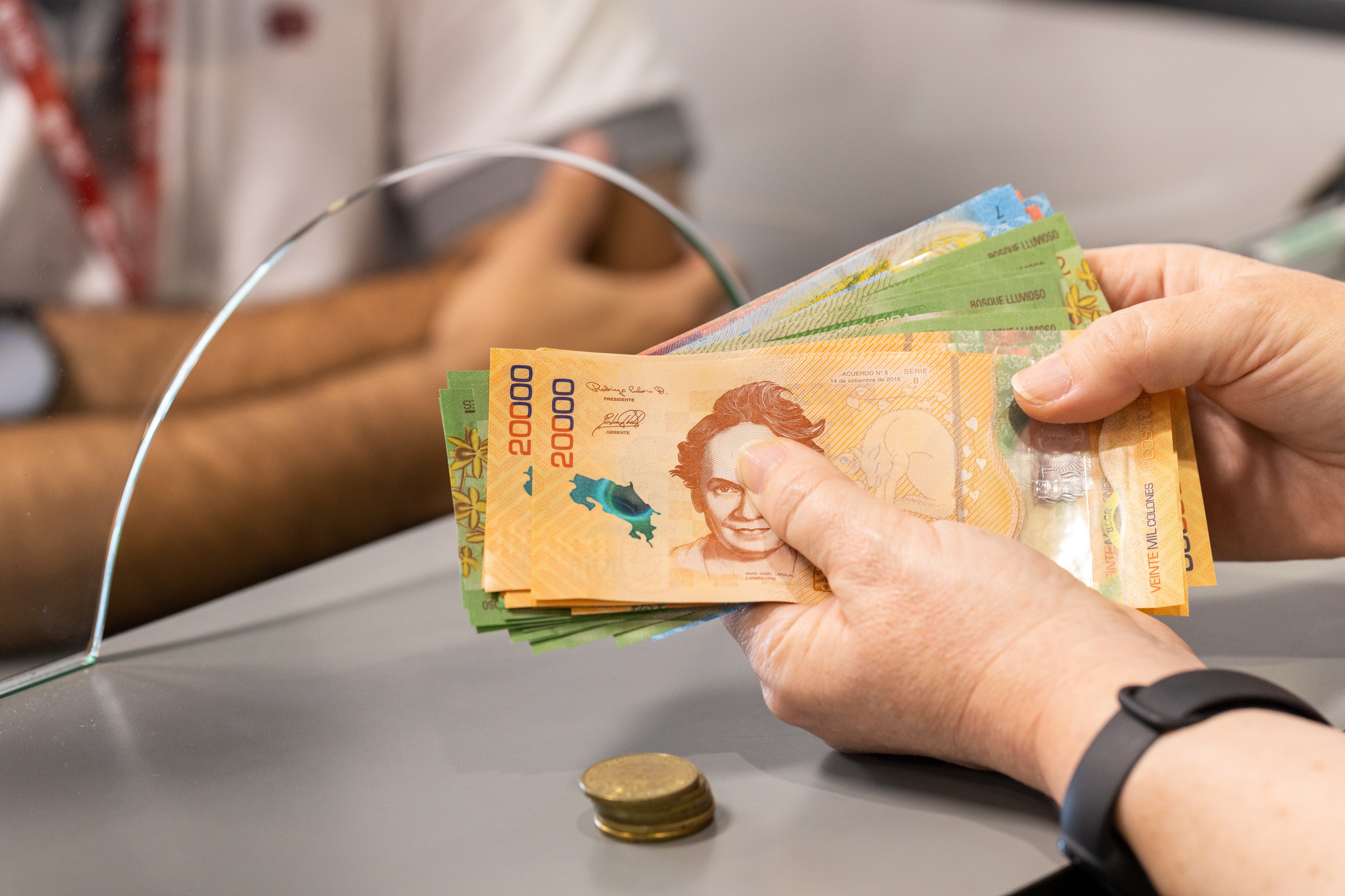 Una mano sostiene un billetes de ¢20.000 colones con una ventanilla de fondo