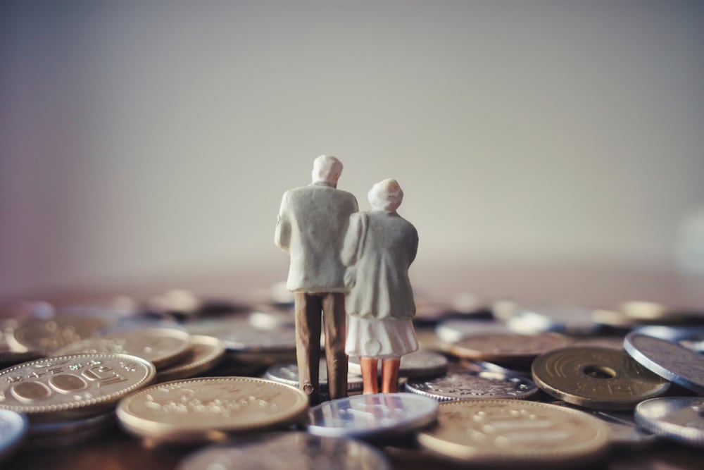 Figura en miniatura de una pareja de adultos mayores de espaldas, de pie sobre un fondo de monedas, representando el sistema de pensiones y la postergación de los fondos generacionales del ROP en Costa Rica.