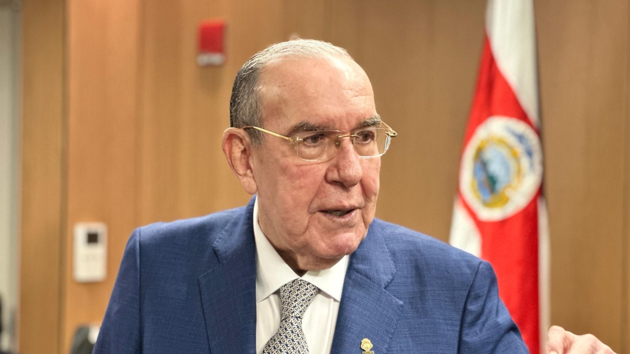 Rodrigo Arias, presidente de la Asamblea Legislativa de Costa Rica, en traje azul, con la bandera nacional al fondo, durante un evento público, en el contexto de críticas recibidas del presidente Rodrigo Chaves.