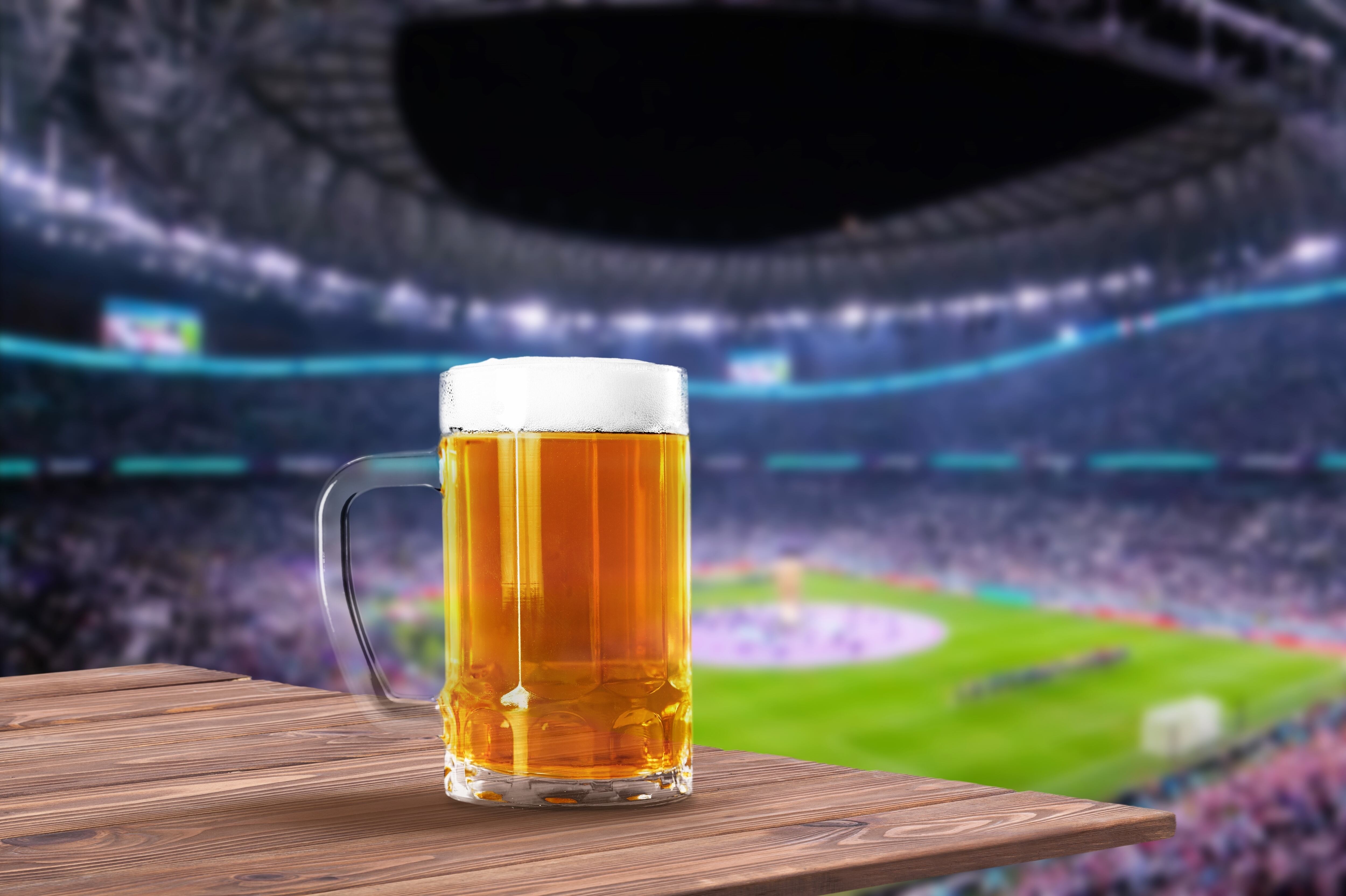 Jarra de cerveza espumante con fondo de estadio, cancha, público en graderías