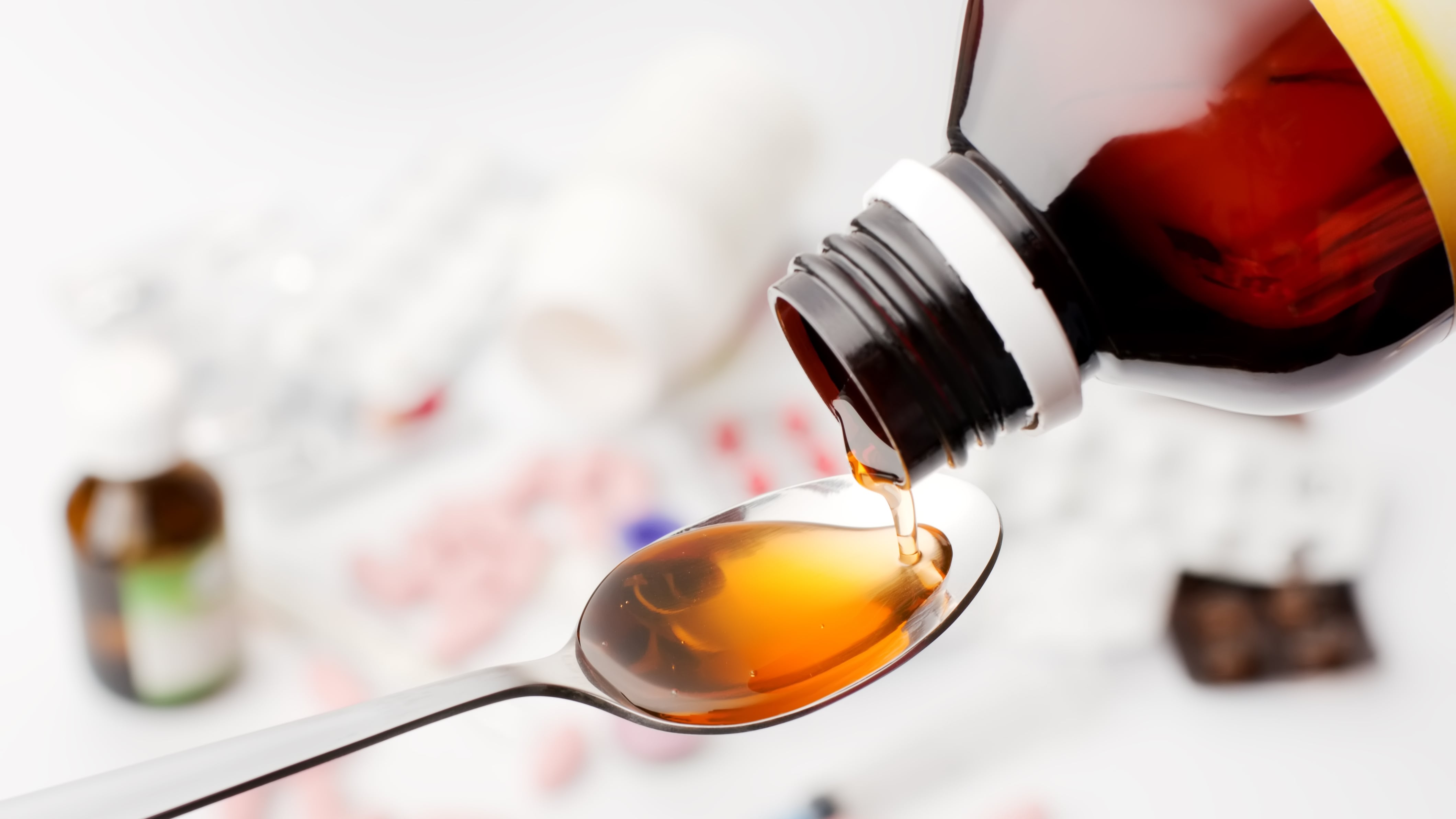 En primer plano: un jarabe sirviéndose en una cuchara. De fondo, desenfocados, varios otros medicamentos.