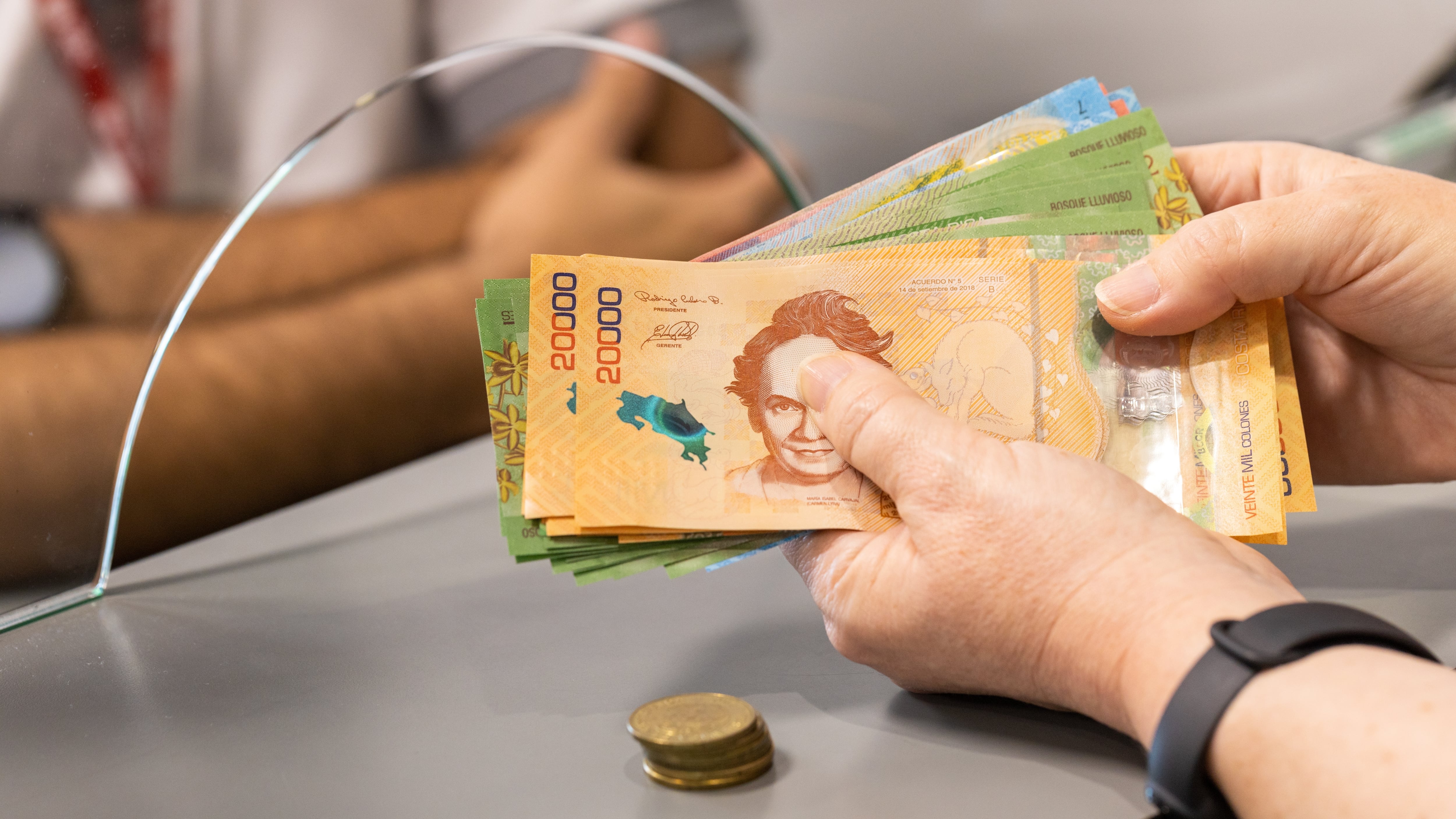 Manos de una persona contando billetes de 20.000 colones en un banco en Costa Rica, con un empleado detrás de un vidrio de atención.