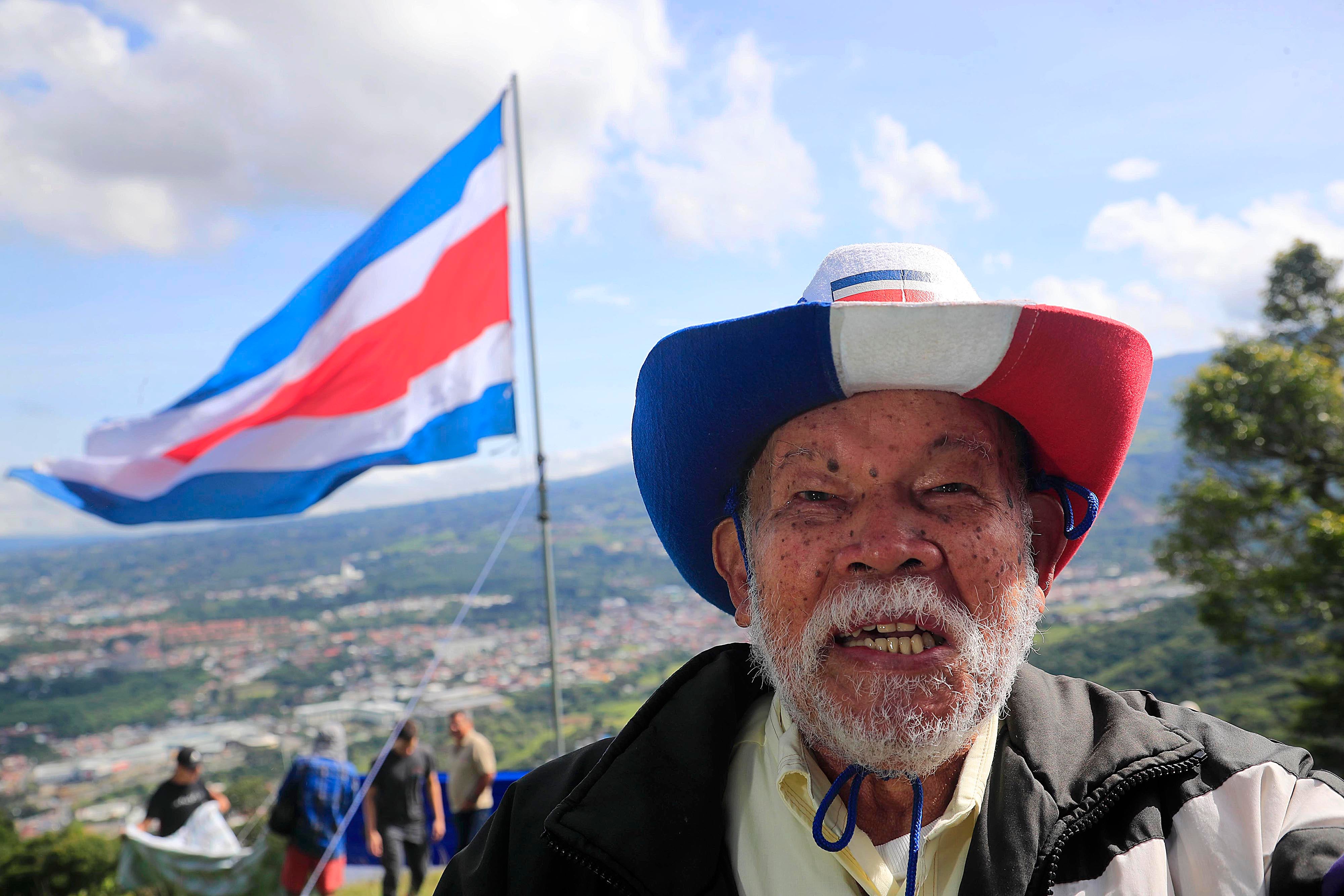 15/09/2023 Tres Ríos. Familiares y amigos de Rigoberto Vega Alvarado izaron una bandera gigante, como ha sido tradición desde hace 60 años, cada 15 de setiembre, en la ladera del potrero La Rosa, en la fila de los cerros La Carpintera. Desde las 5:00 a.m. salieron del centro del cantón de La Unión unas 30 personas, bien equipadas con herramientas, telas, tubos, cuerdas, y por supuesto la bandera. Y ya en el sitio empezaron a colocar las seis franjas de tela horizontales de 14 metros por 1 y medio de alto a una distancia de 10 metros aproximadamente cada una, de manera que desde el centro de Tres Ríos se aprecia como una sola bandera por efecto de la aproximación de los planos. Todos trabajan porque mientras unos se ocupan de colocar las franjas individuales de tela, otros trabajan en la colocación del tubo gigante para luego izar la bandera, y otros más, especialmente las mujeres se ocupan de hacer el café, la aguadulce y los sandwiches. Mientras se trabaja en el levantamiento de la bandera y la colocación de las telas individuales las solo el viento compite con las notas de canciones tradicionales del folklor costarricense propias de la época que salen de un enorme parlante.