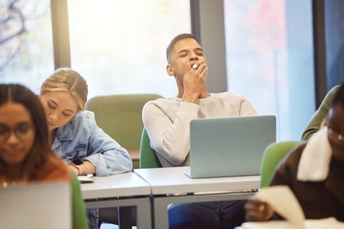 Estudiantes en un aula universitaria, uno de ellos bostezando frente a su laptop, simbolizando el impacto de los desafíos culturales, económicos y familiares en la juventud.