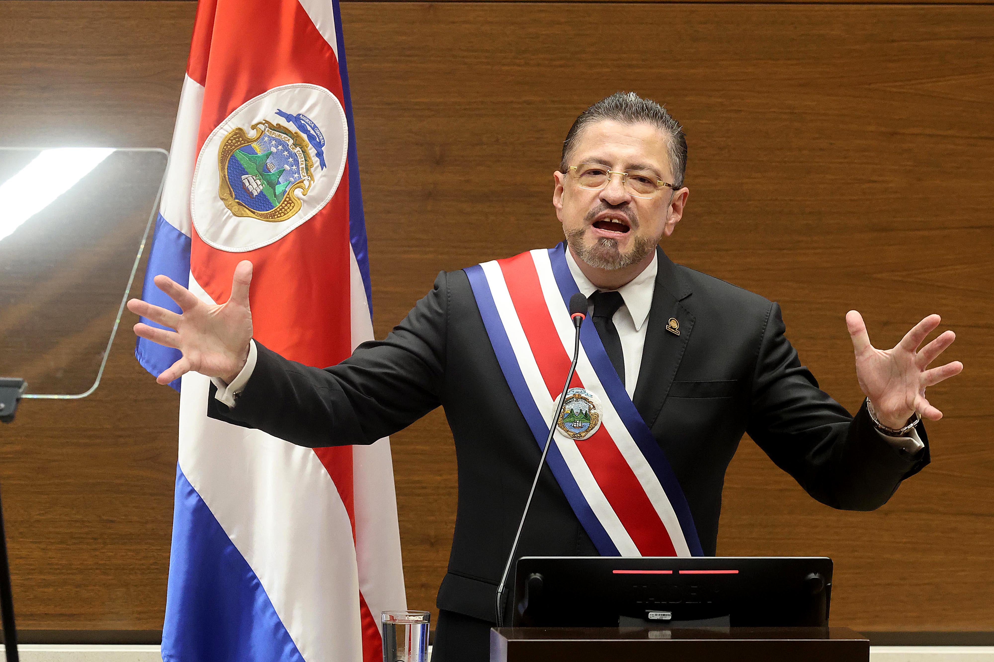Retrato de un presidente vestido con traje entero oscuro y banda presidencial con la bandera de Costa Rica. De pie en un podio, gesticula en un discurso.