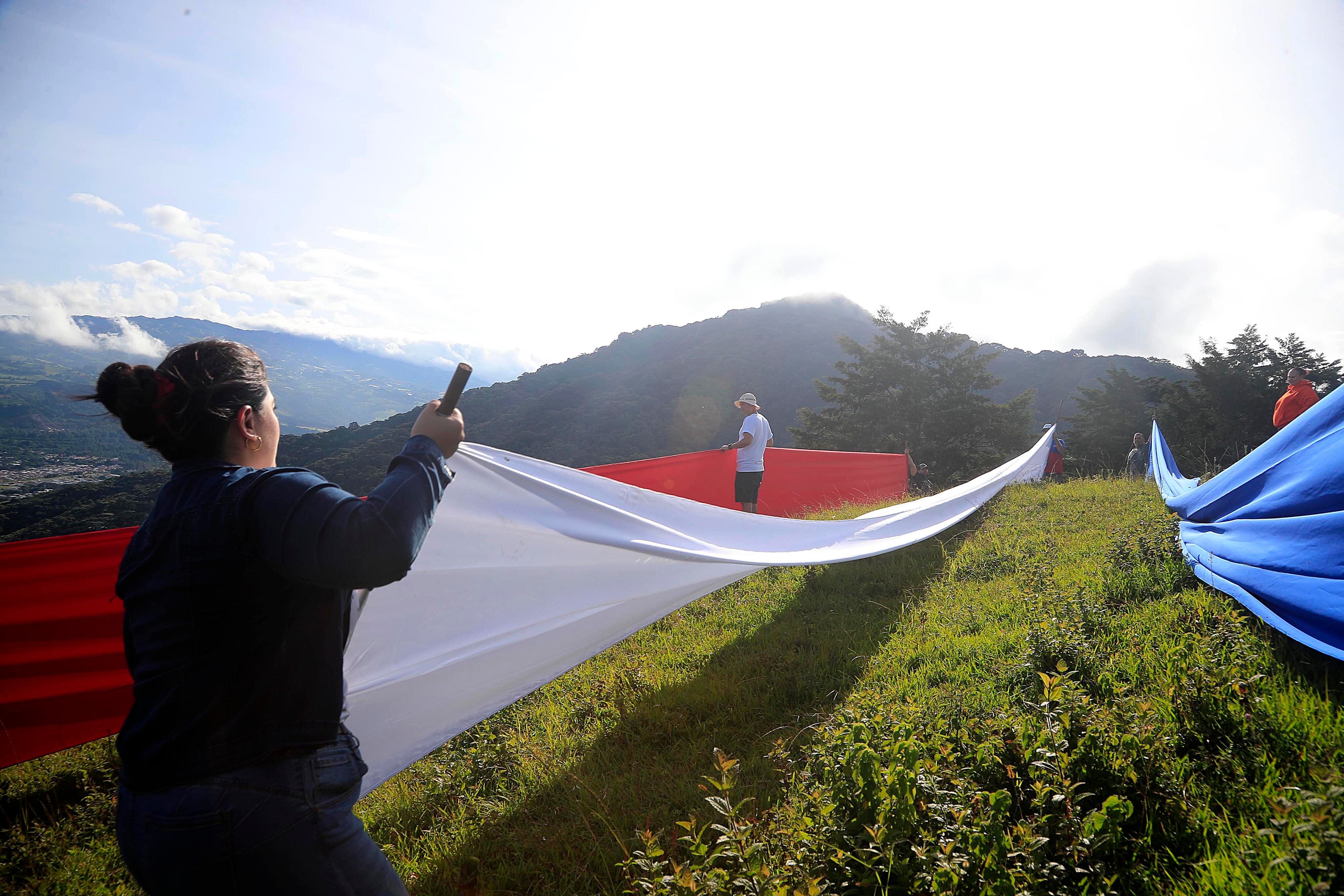 15/09/2023 Tres Ríos. Familiares y amigos de Rigoberto Vega Alvarado izaron una bandera gigante, como ha sido tradición desde hace 60 años, cada 15 de setiembre, en la ladera del potrero La Rosa, en la fila de los cerros La Carpintera. Desde las 5:00 a.m. salieron del centro del cantón de La Unión unas 30 personas, bien equipadas con herramientas, telas, tubos, cuerdas, y por supuesto la bandera. Y ya en el sitio empezaron a colocar las seis franjas de tela horizontales de 14 metros por 1 y medio de alto a una distancia de 10 metros aproximadamente cada una, de manera que desde el centro de Tres Ríos se aprecia como una sola bandera por efecto de la aproximación de los planos. Todos trabajan porque mientras unos se ocupan de colocar las franjas individuales de tela, otros trabajan en la colocación del tubo gigante para luego izar la bandera, y otros más, especialmente las mujeres se ocupan de hacer el café, la aguadulce y los sandwiches. Mientras se trabaja en el levantamiento de la bandera y la colocación de las telas individuales las solo el viento compite con las notas de canciones tradicionales del folklor costarricense propias de la época que salen de un enorme parlante.