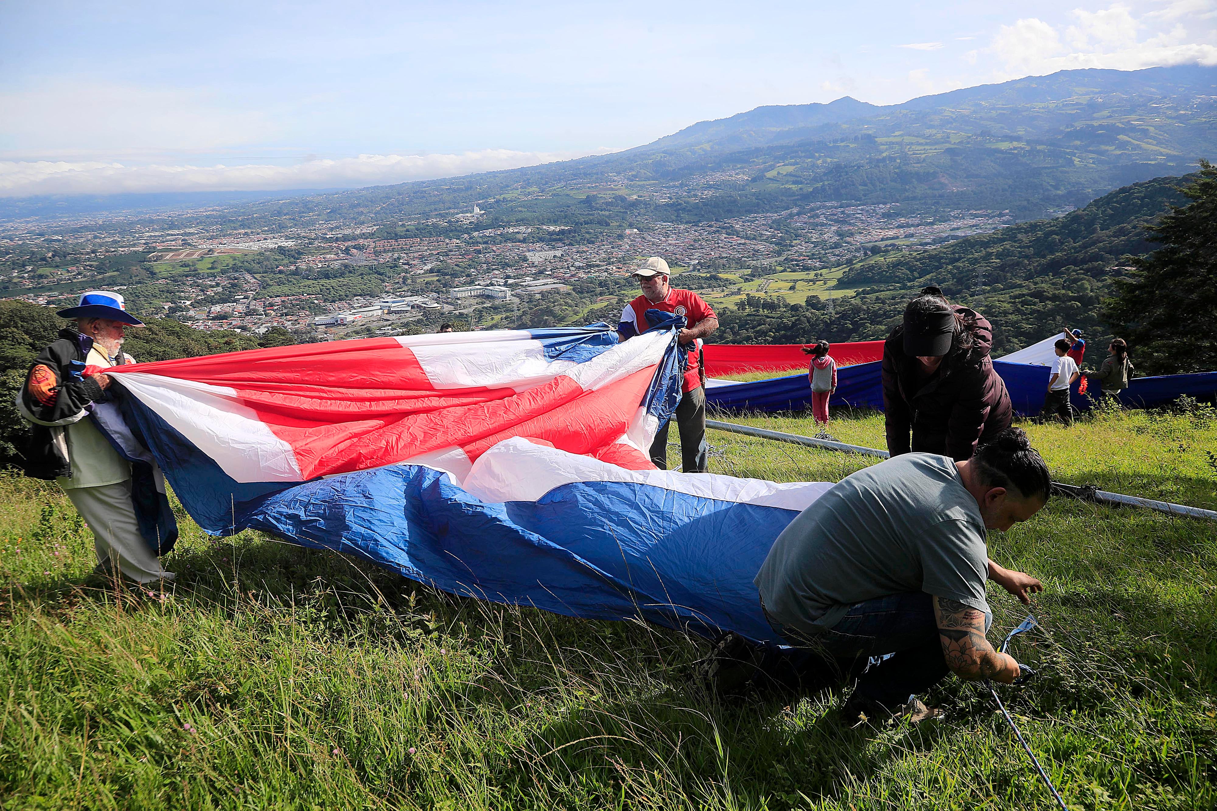 15/09/2023 Tres Ríos. Familiares y amigos de Rigoberto Vega Alvarado izaron una bandera gigante, como ha sido tradición desde hace 60 años, cada 15 de setiembre, en la ladera del potrero La Rosa, en la fila de los cerros La Carpintera. Desde las 5:00 a.m. salieron del centro del cantón de La Unión unas 30 personas, bien equipadas con herramientas, telas, tubos, cuerdas, y por supuesto la bandera. Y ya en el sitio empezaron a colocar las seis franjas de tela horizontales de 14 metros por 1 y medio de alto a una distancia de 10 metros aproximadamente cada una, de manera que desde el centro de Tres Ríos se aprecia como una sola bandera por efecto de la aproximación de los planos. Todos trabajan porque mientras unos se ocupan de colocar las franjas individuales de tela, otros trabajan en la colocación del tubo gigante para luego izar la bandera, y otros más, especialmente las mujeres se ocupan de hacer el café, la aguadulce y los sandwiches. Mientras se trabaja en el levantamiento de la bandera y la colocación de las telas individuales las solo el viento compite con las notas de canciones tradicionales del folklor costarricense propias de la época que salen de un enorme parlante.