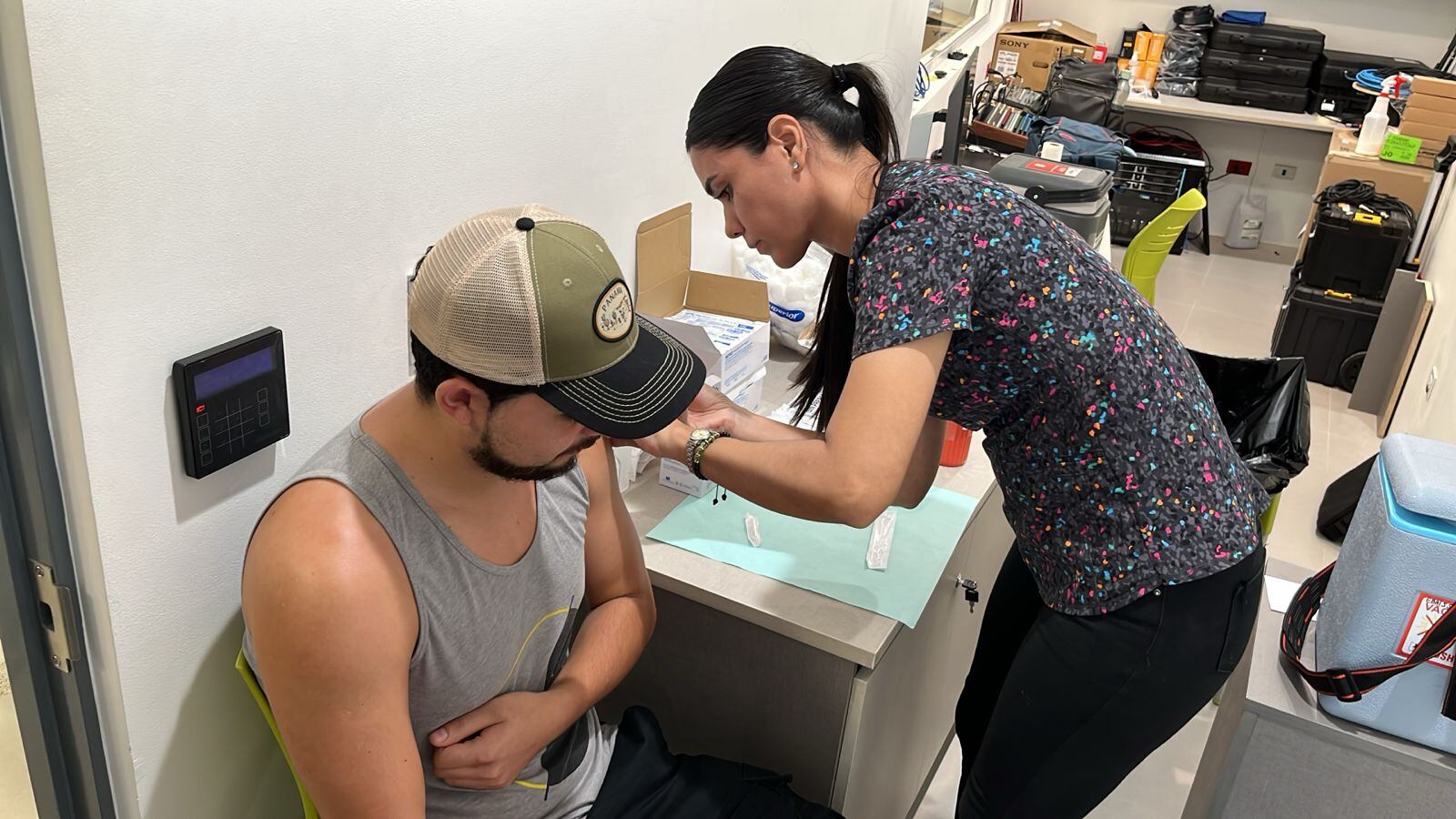 Mujer vacuna a paciente masculino en su brazo izquierdo. Ella, de pie. Él, sentado, con camiseta de tirantes y gorra.