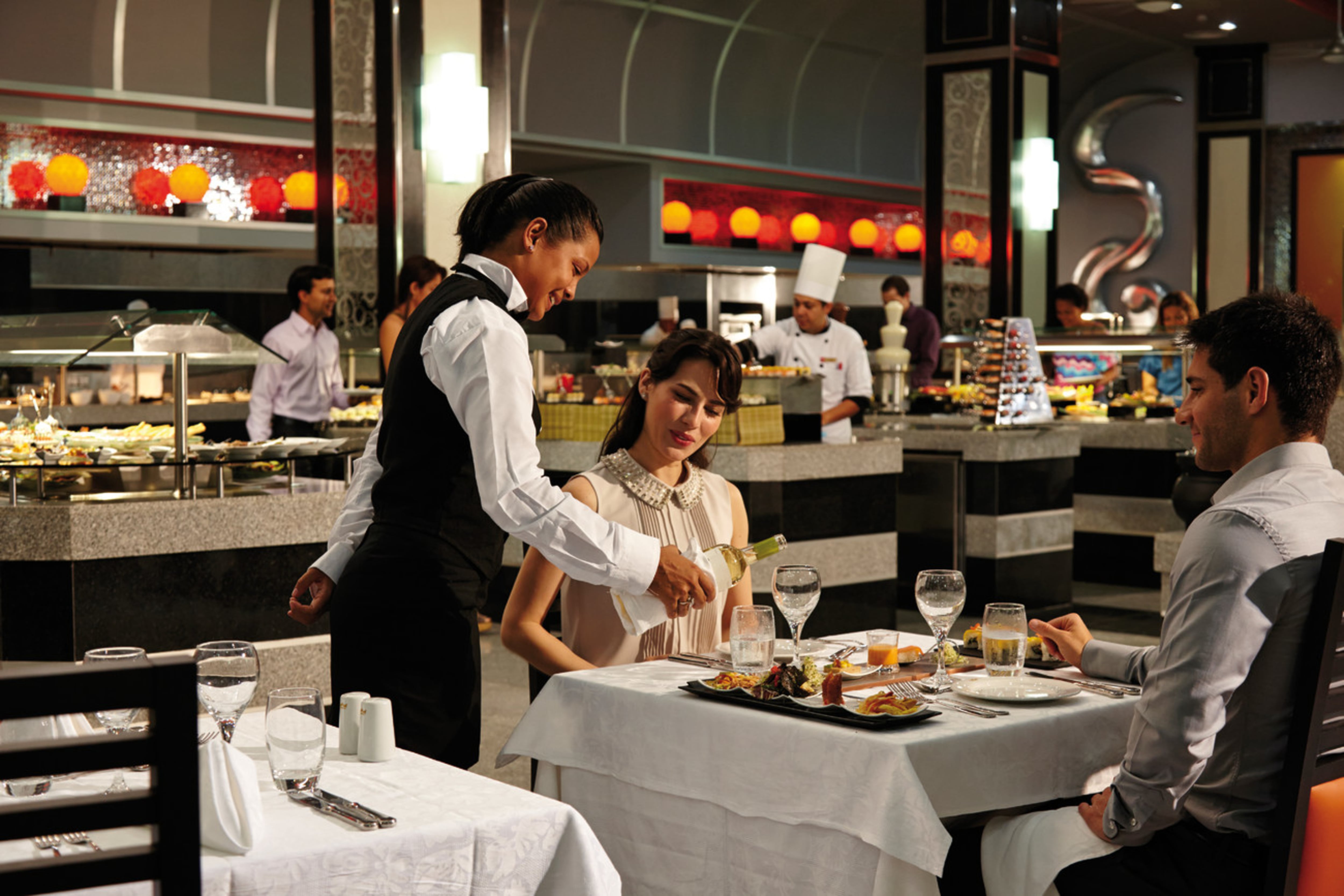 Una camarera sirve vino a una pareja en el restaurante de un hotel todo incluido en Costa Rica, con un fondo decorado y un buffet. La escena destaca el ambiente acogedor y el servicio personalizado del modelo todo incluido.