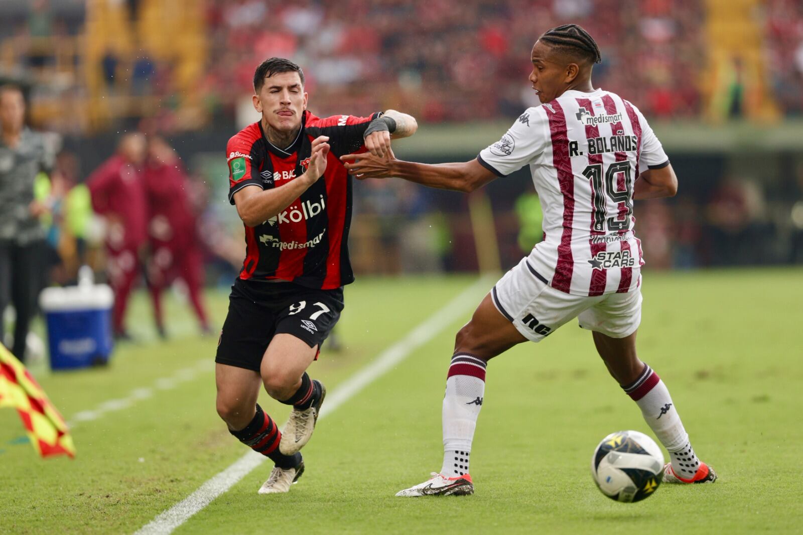 Dos jugadores, uno de Alajuelense con uniforme rojinegro, llamado Anderson Canhoto, y otro de Saprissa con uniforme blanco con franjas moradas, llamado Ryan Bolaños, disputan un balón en un partido de fútbol.