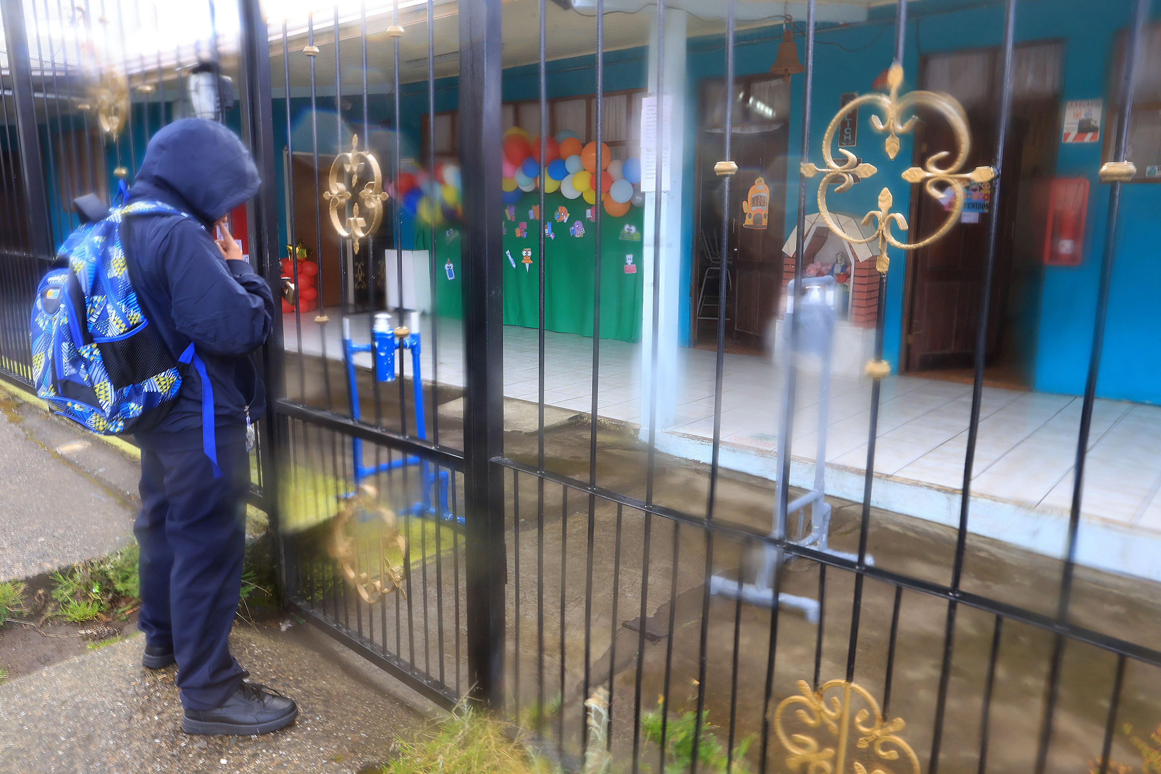 Niño con bulto en uniforme frente a reja de escuela, al fondo las aulas.