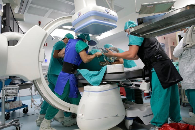 En un quirófano, varias personas vestidas de verde sostienen a un paciente para hacerle un examen con un aparato con forma de arco en C.