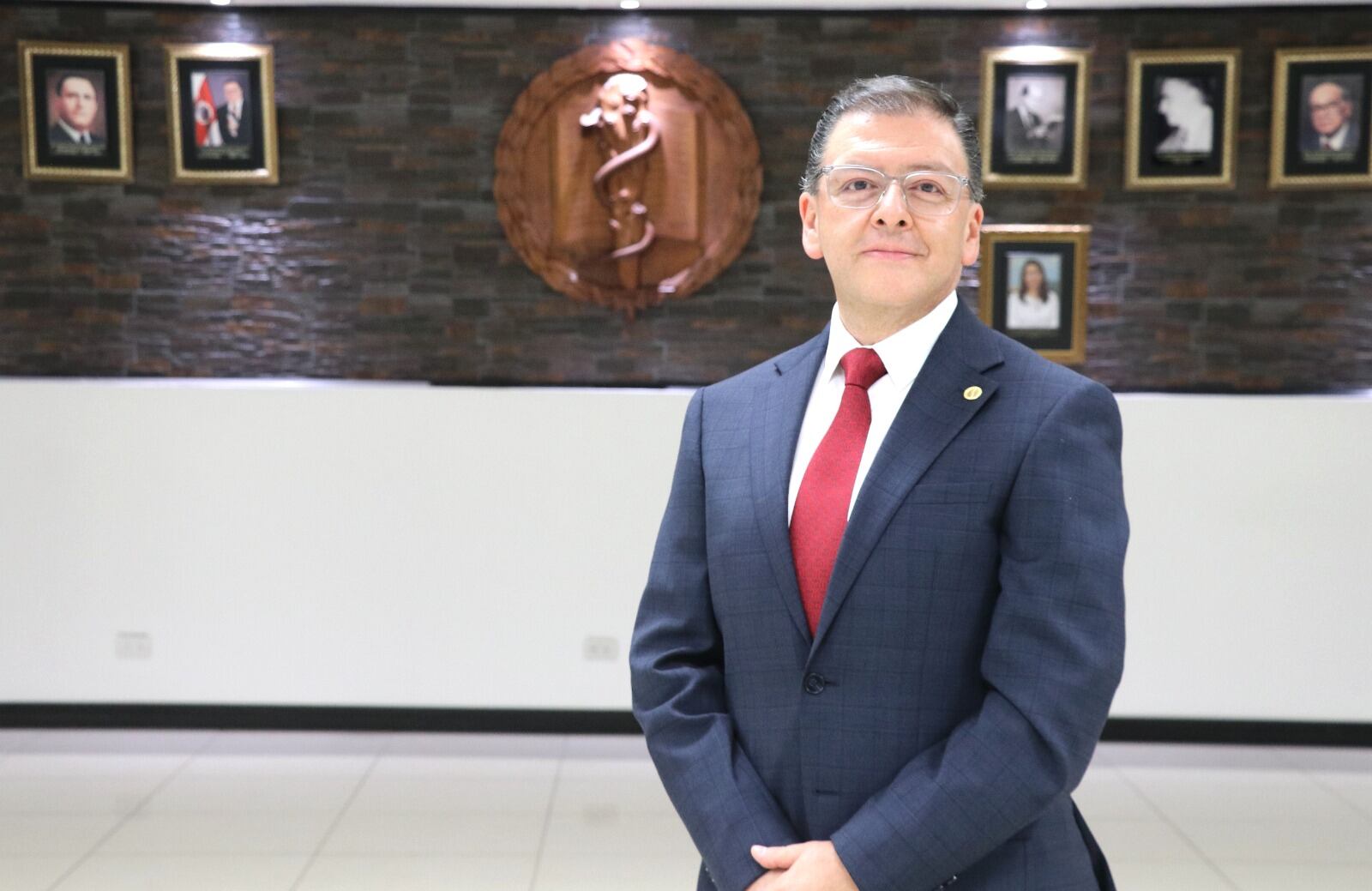 Retrato de hombre de mediana edad, blanco, delgado, con traje entero azul, corbata roja, manos enlazadas al frente.