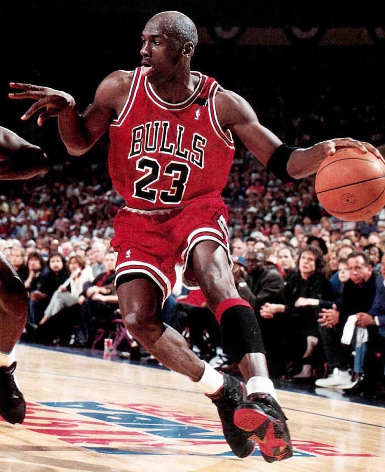 Imagen de Michael Jordan cuando jugaba con los Bulls de Chicago, usa su conocido uniforme de color rojo, pica el balón y tiene la lengua afuera.