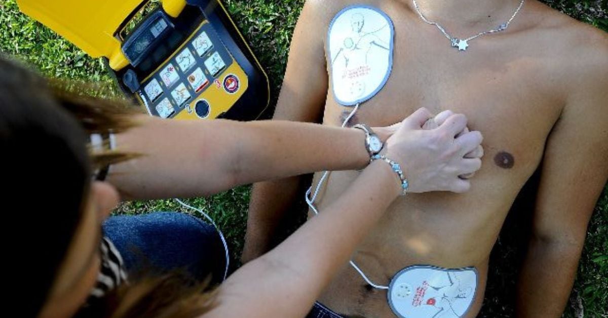 Una persona utiliza un desfibrilador externo automático (DEA) en un paciente durante una maniobra de resucitación en un área verde, con un dispositivo amarillo visible junto al afectado.