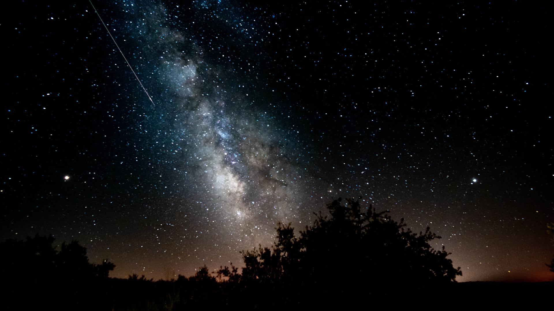 Fotografía de larga exposición de rastros estelares cruzando el cielo nocturno sobre un paisaje montañoso con un lago en primer plano.