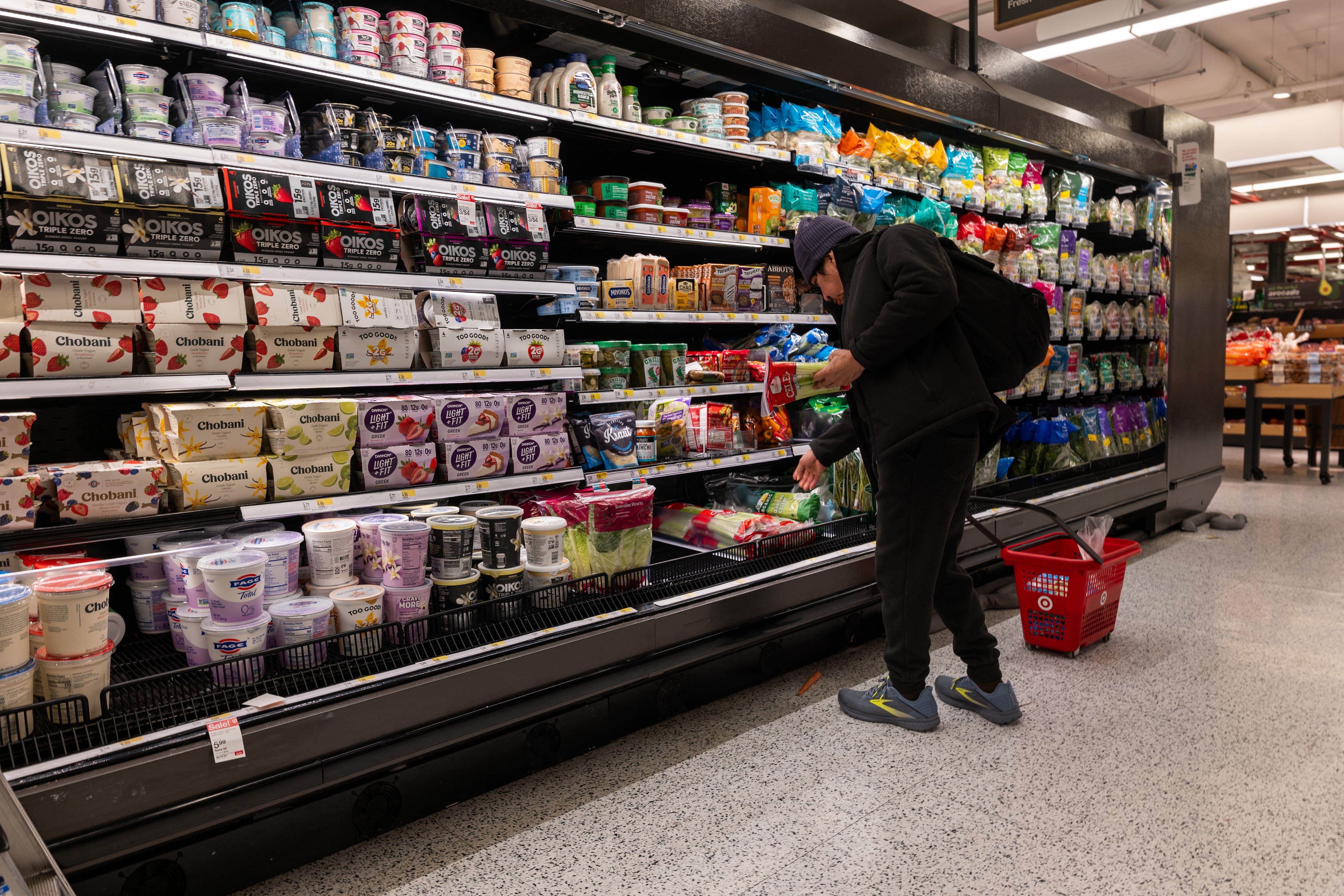 Un cliente revisando productos en el área de refrigerados de un supermercado en Estados Unidos, con una canasta roja en el suelo.