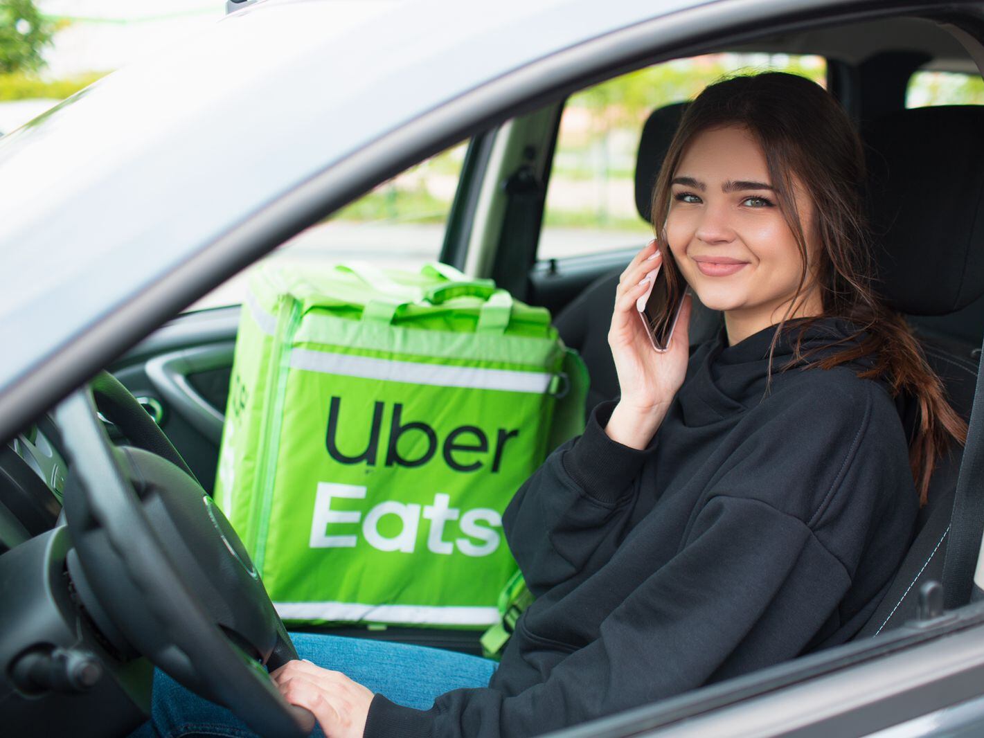Foto ilustrativa de una mujer sonriente al volante, con su celular en la oreja y un bolso de Uber Eats en el asiento de copiloto