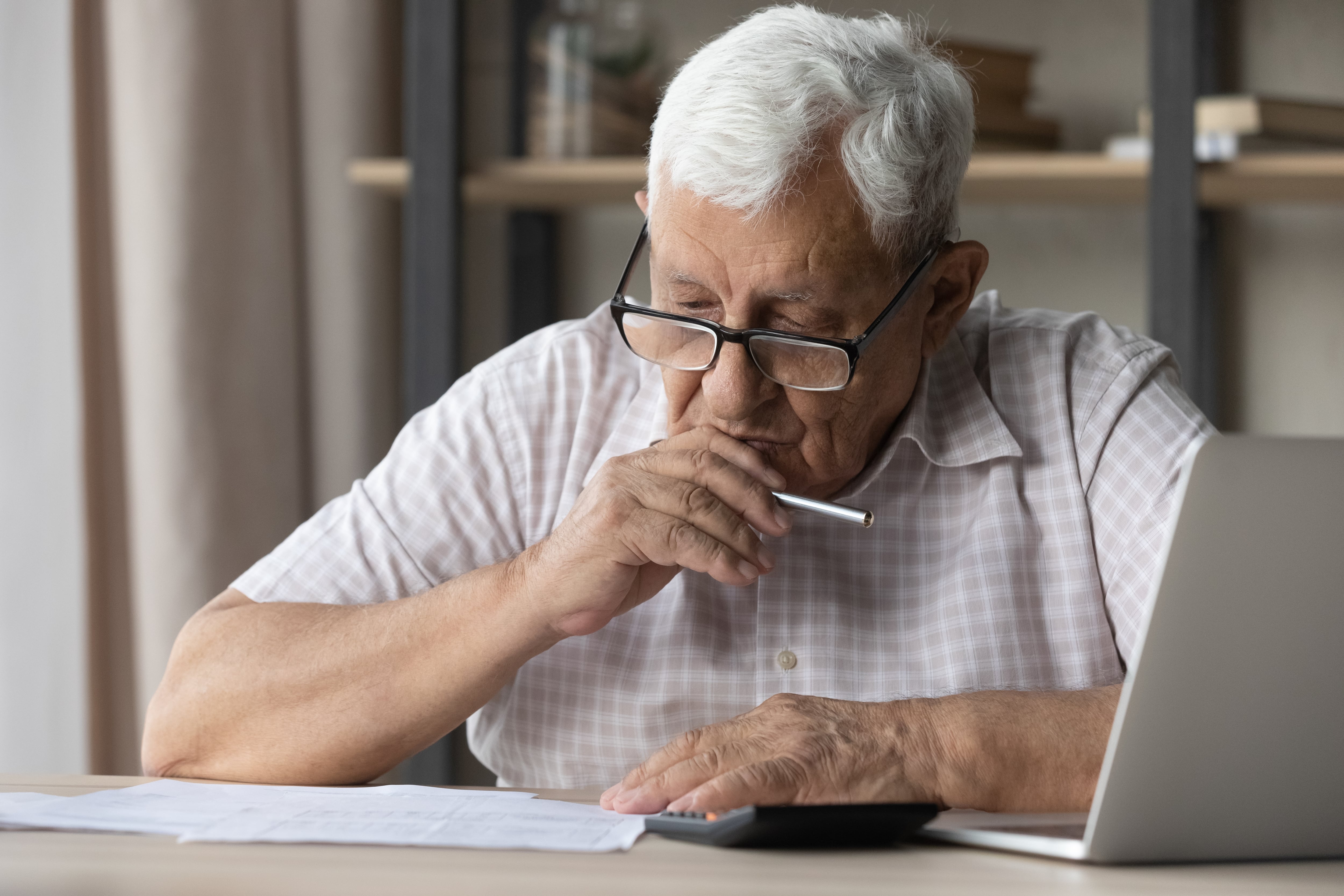 Adulto mayor viendo sus cuentas, papel y calculadora en mano, y a un lado la laptop, computadora