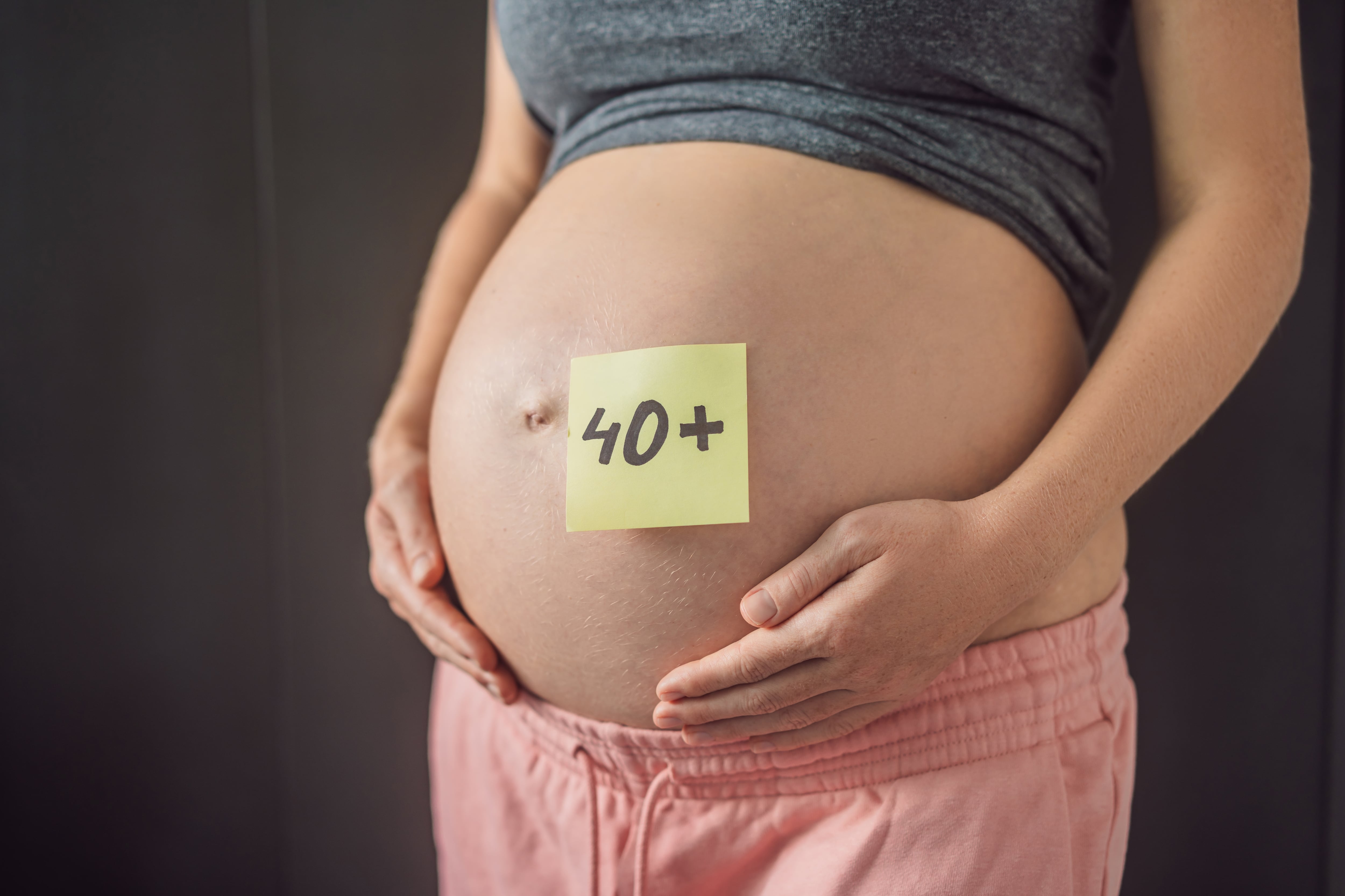 Una mujer embarazada, en su vientre a un papel con la frase 40+