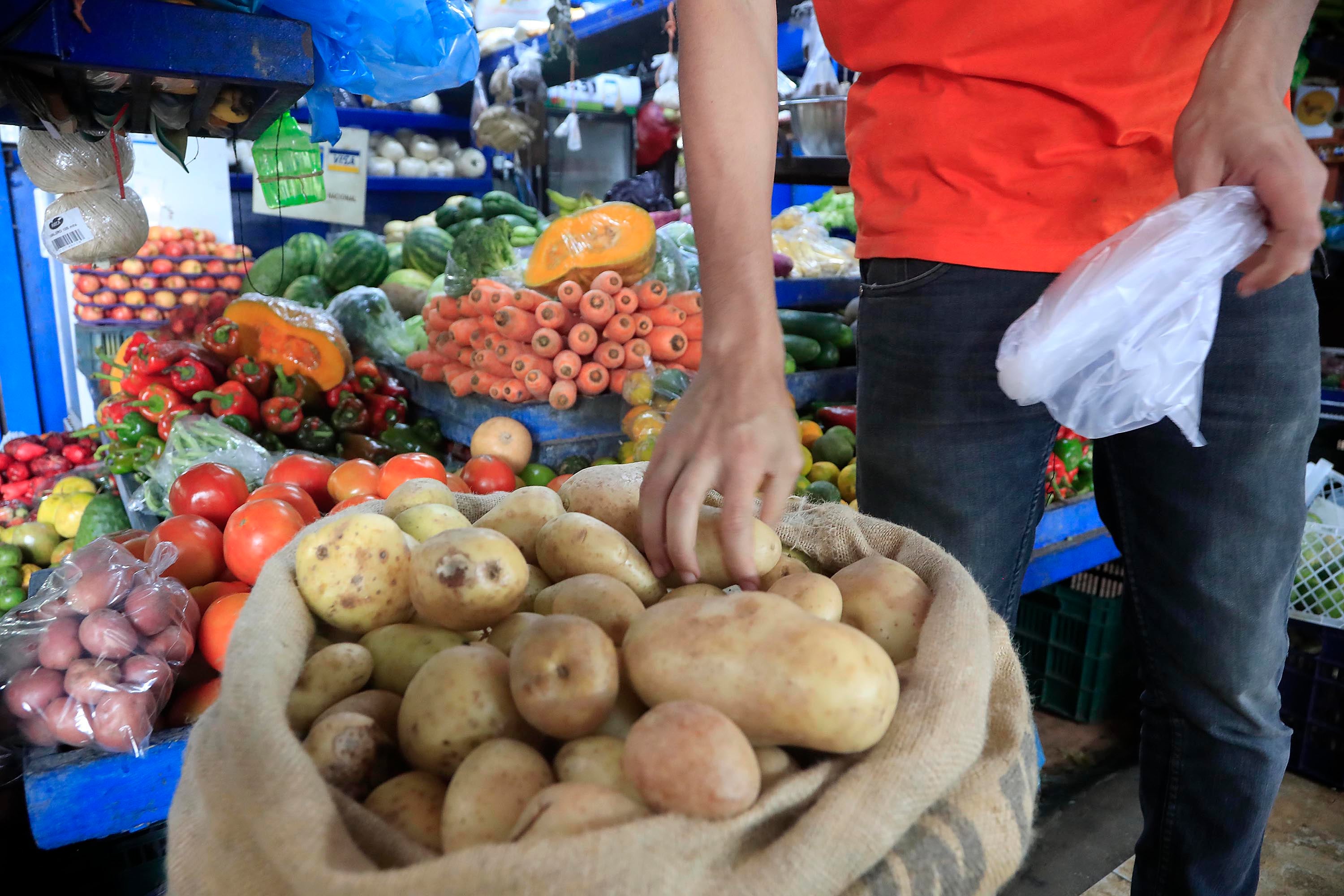 Un cliente escoge papas de un saco en un mercado, rodeado de vegetales como zanahorias, tomates y chiles dulces, ilustrando el impacto del aumento en los precios de alimentos frescos en Costa Rica durante 2024.