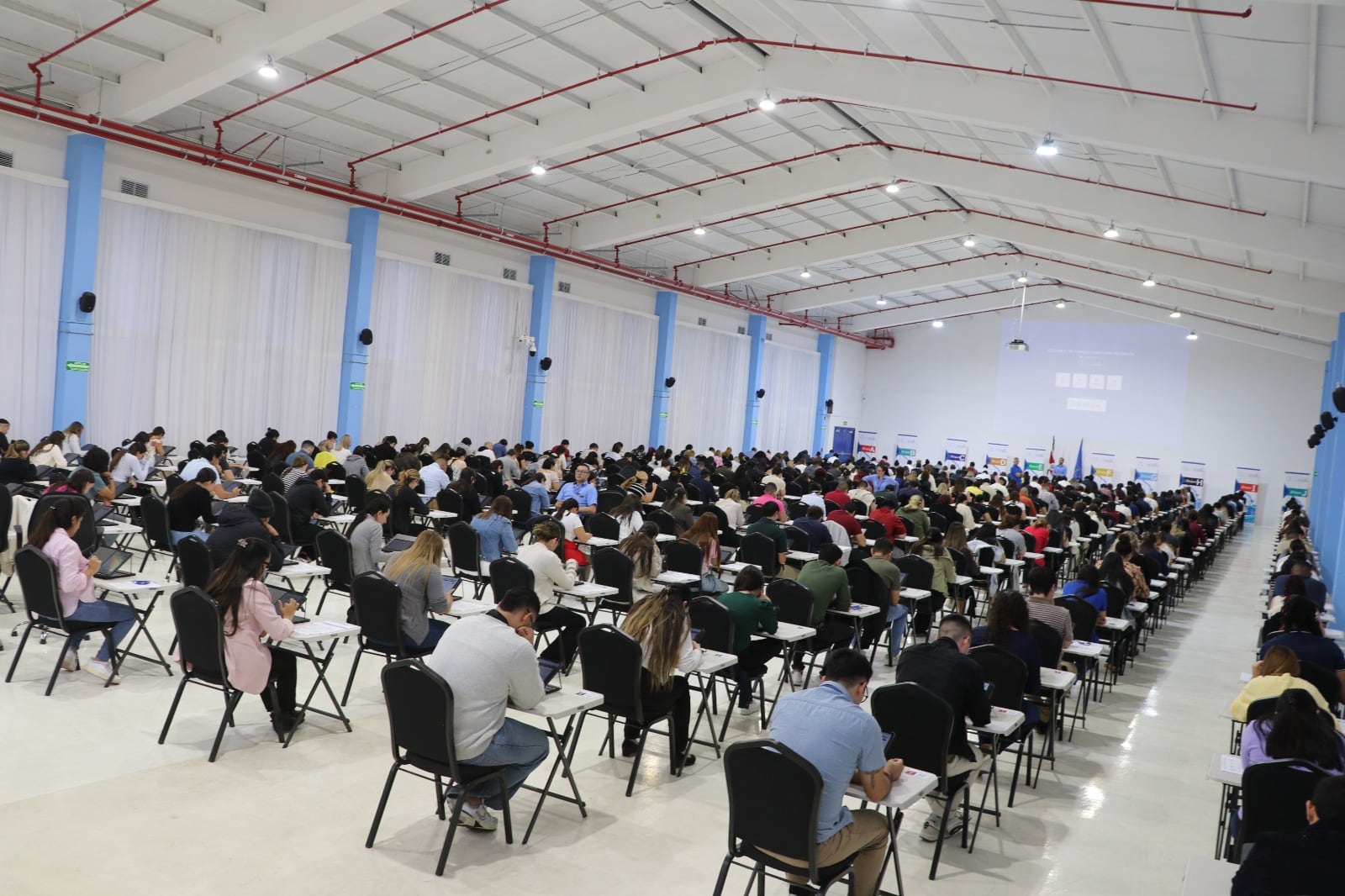 Un grupo de personas sentadas en escritoros realizando un examen en un auditorio. Todas se ven de espaldas.