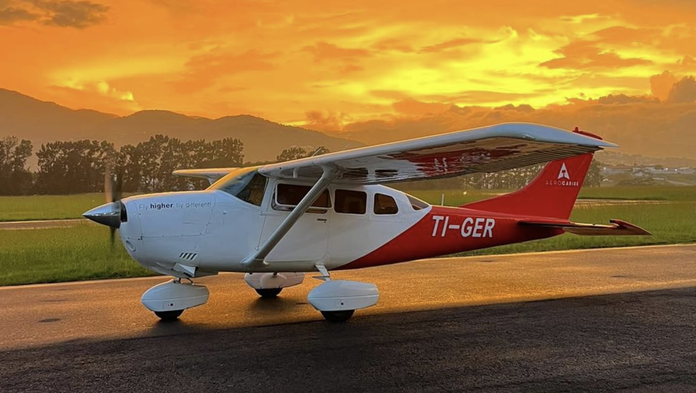 Avioneta Cessna U206G matrícula TI-GER en un aeropuerto al atardecer, con un cielo naranja y montañas al fondo.