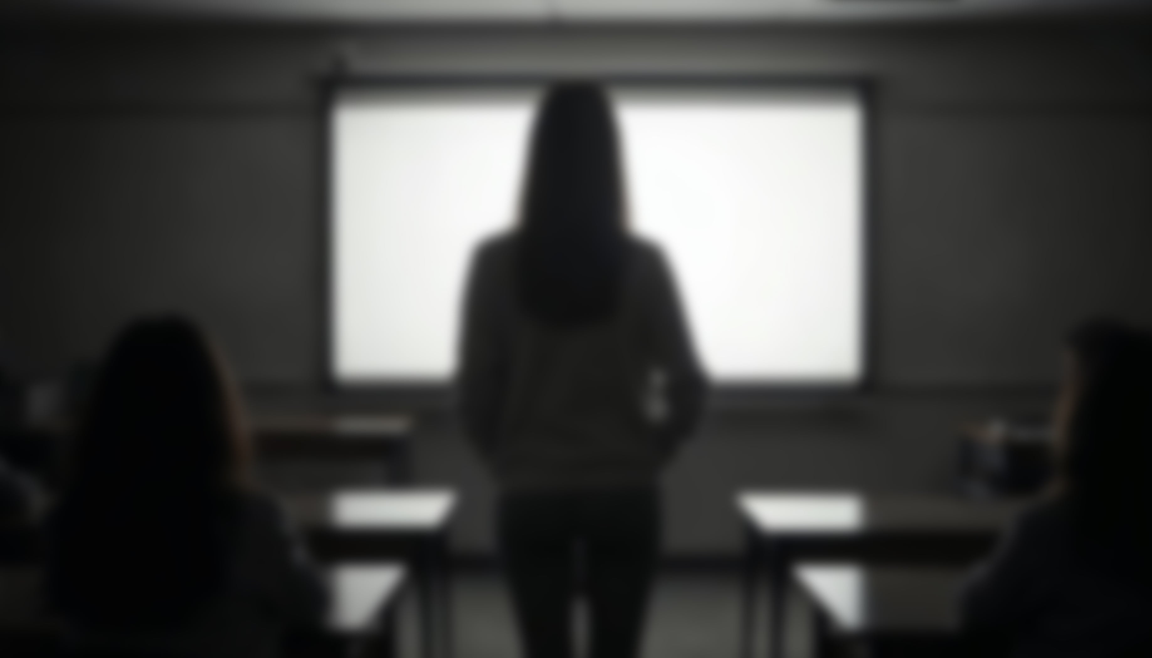 Lado oscuro de la educación. Imagen borrosa, desenfocada y oscura de maestra, profesora, docente de espaldas, en aula con pupitres y una pizarra iluminada al frente