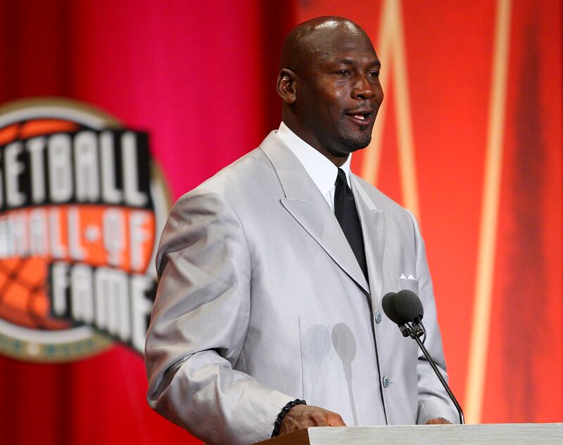 Michael Jordan hablando al micrófono, usa un traje de color gris, camisa blanca y corbata negra.