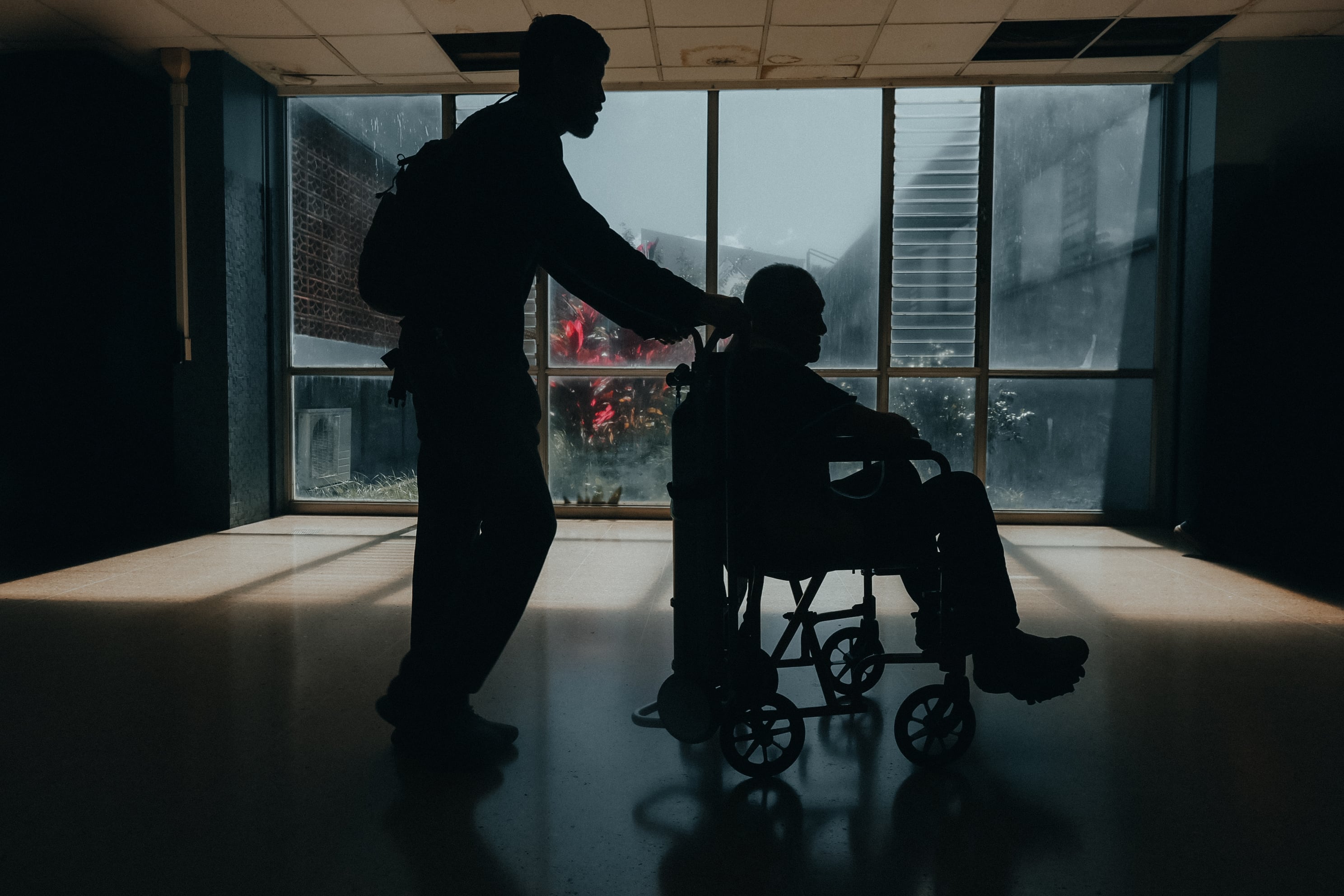 Un adulto mayor es llevado en silla de ruedas por un joven. La imagen está a contraluz. De fondo, se observa una ventana que da a un jardín, en el servicio de consulta externa del Hospital México.