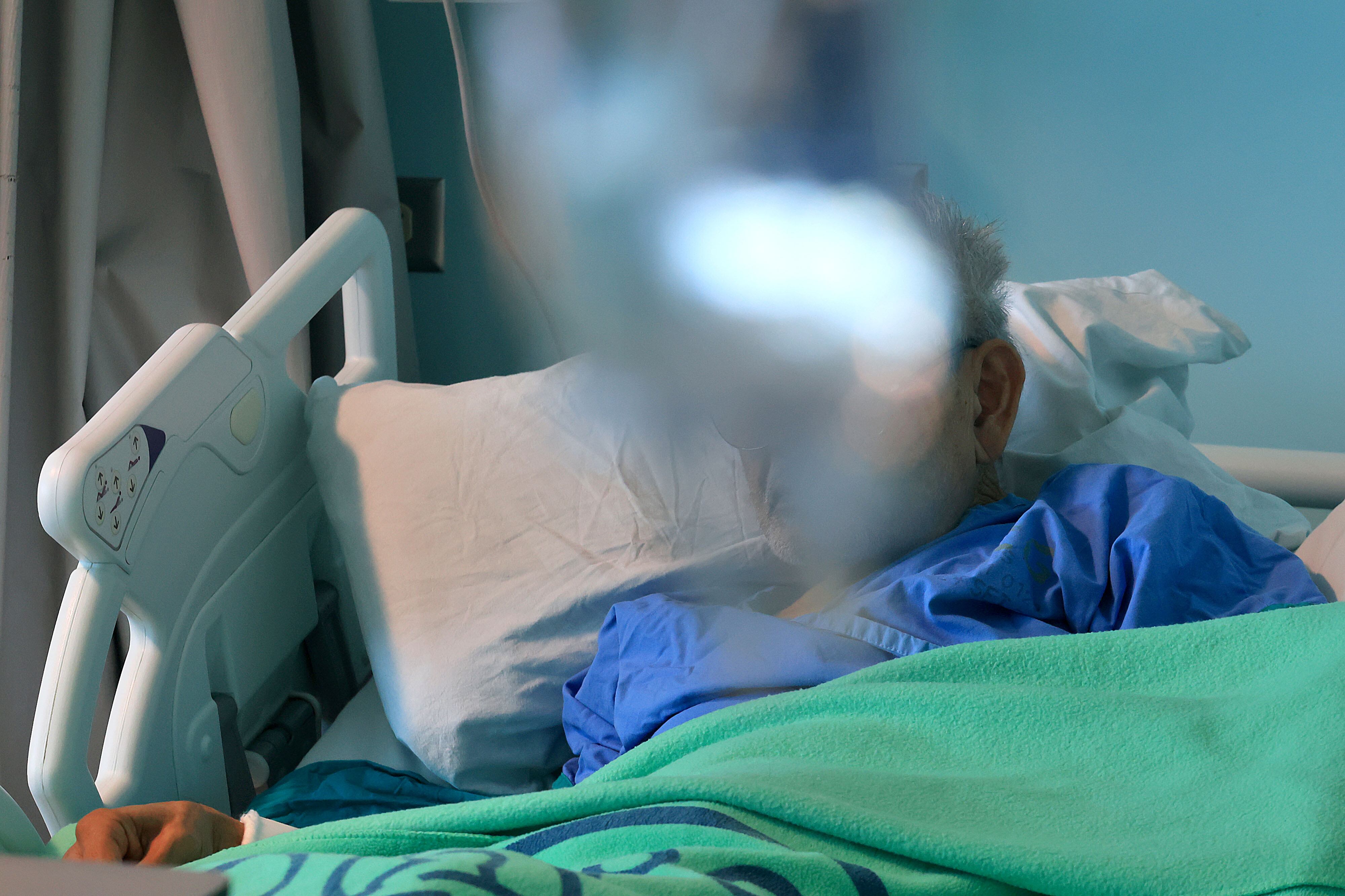 Persona con rostro difuminado. Acostada en una cama de hospital, cubierta con cobija verde. Al fondo, pared celeste.