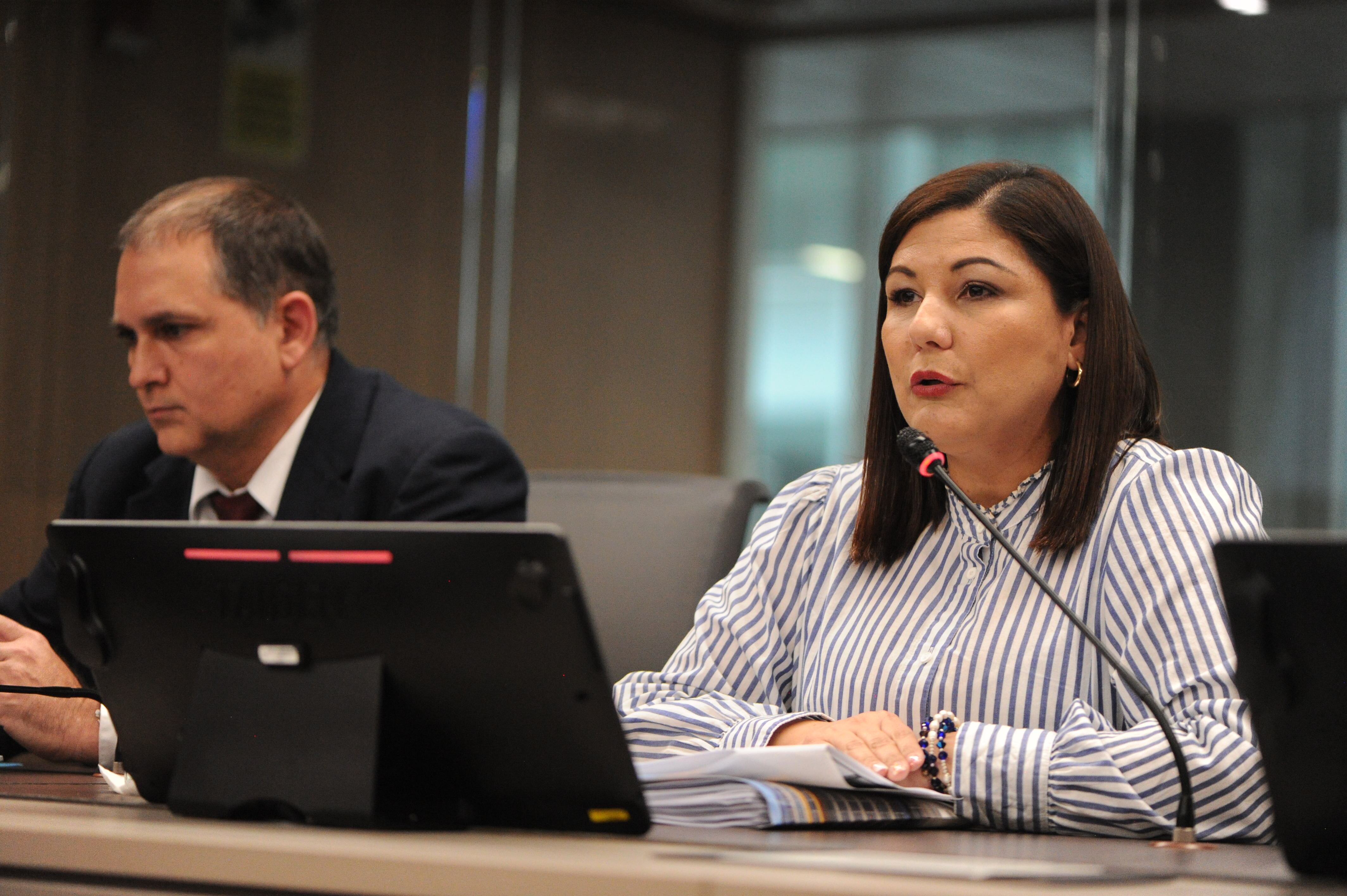 Mujer joven, pelo lacio hasta los hombros, blusa blanca de rayas. A su lado, hombre mayor, con saco azul.