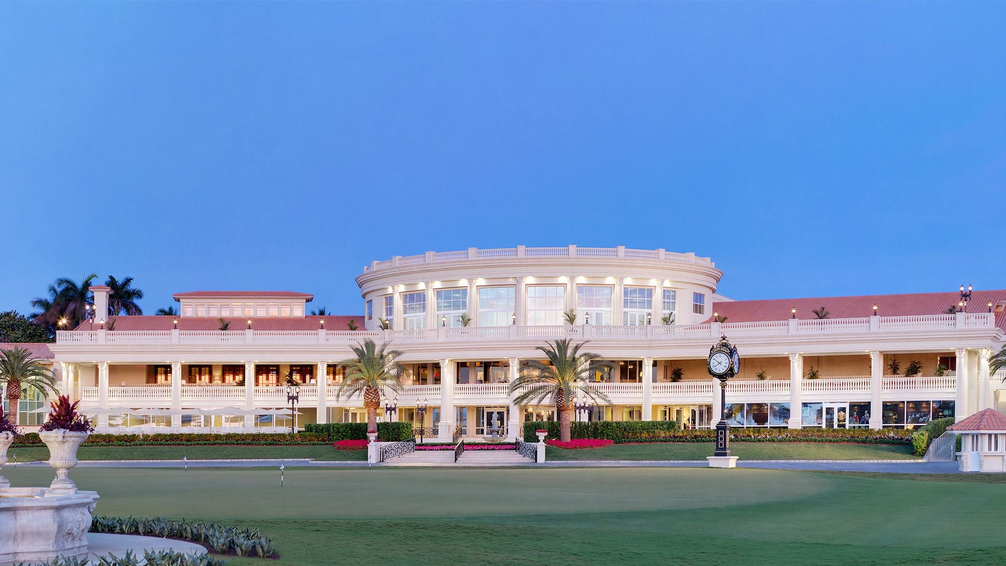 Edificio blanco de estilo clásico con múltiples niveles y balcones, iluminado al anochecer frente a un campo de golf perfectamente cuidado.