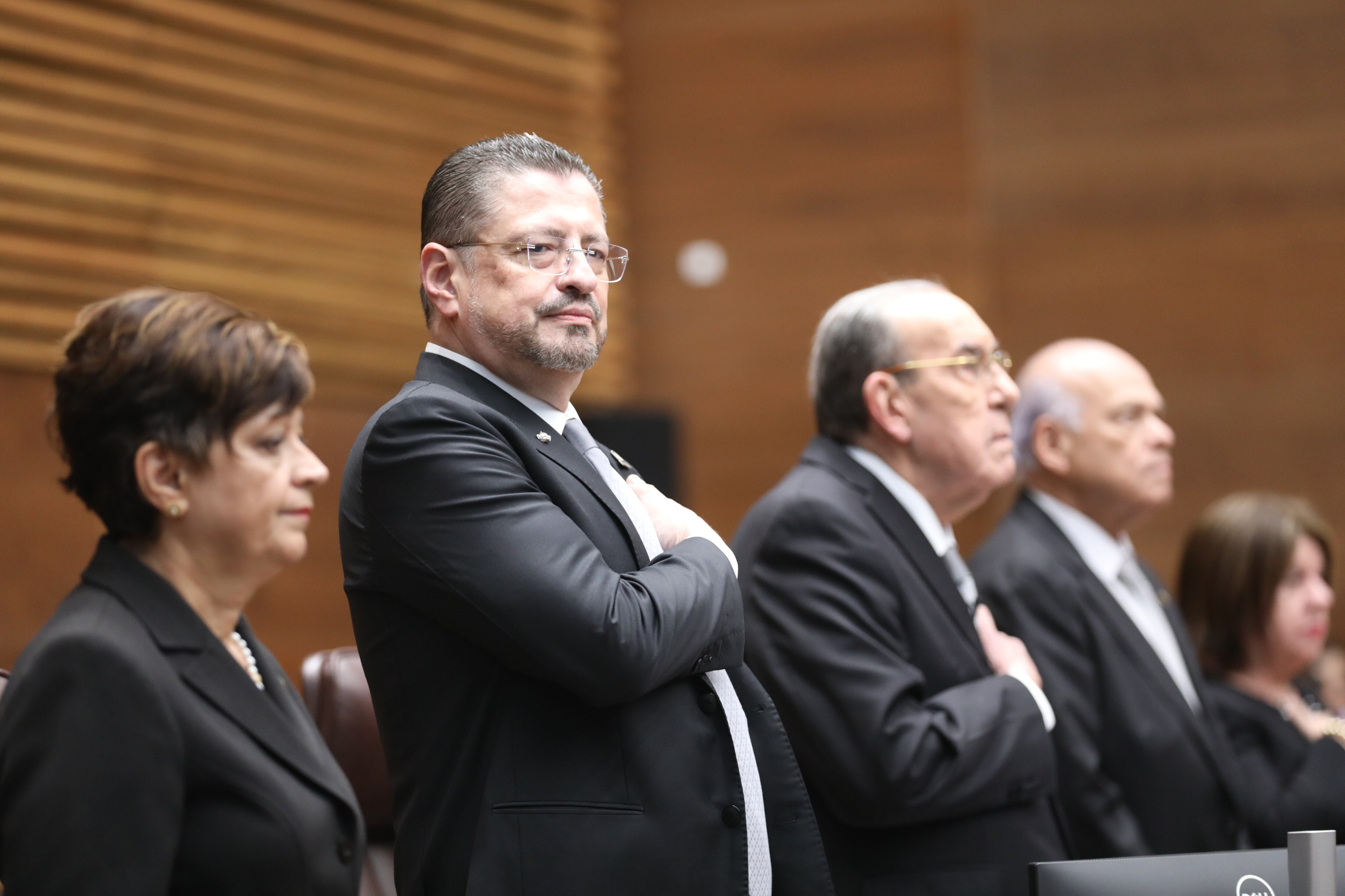 El presidente de Costa Rica, Rodrigo Chaves Robles, de pie con la mano en el pecho en un acto oficial en la Asamblea Legislativa, acompañado por otros funcionarios del Gobierno y la Asamblea Legislativa.