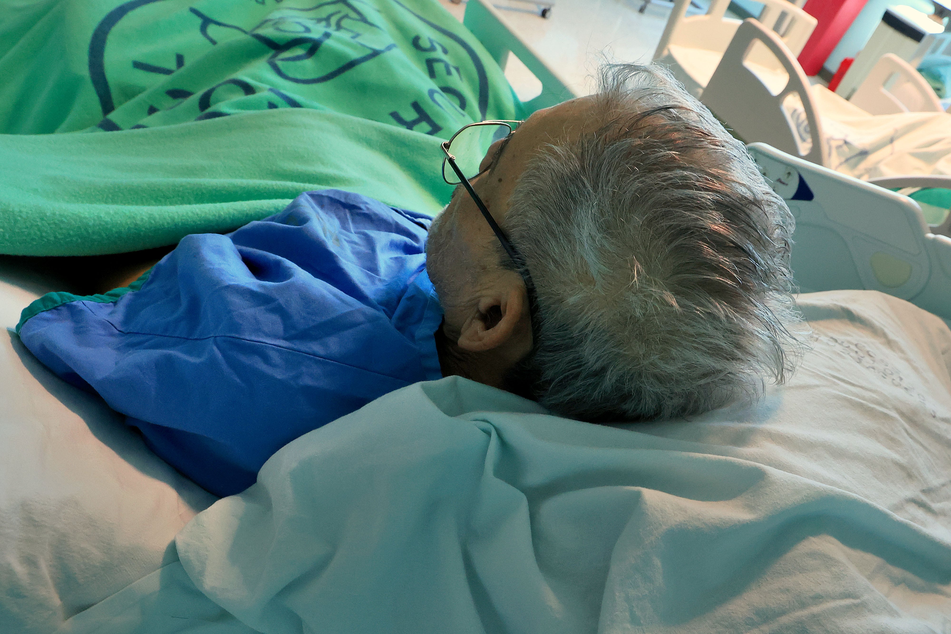 Imagen cenital de adulto mayor acostado en cama de hospital. Pelo blanco, con anteojos.