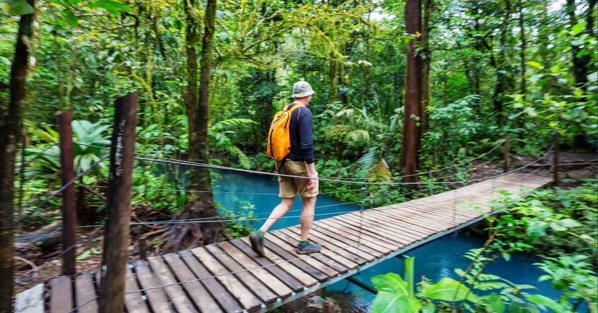 Turista con mochila naranja cruzando un puente colgante de madera sobre el Río Celeste, rodeado de exuberante vegetación en el Parque Nacional Volcán Tenorio, Costa Rica. Destino popular durante Semana Santa.