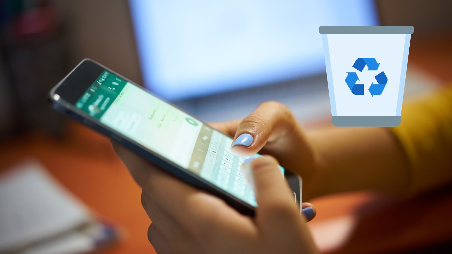 Primer plano de una persona utilizando un celular para eliminar archivos de WhatsApp con el fin de liberar espacio de almacenamiento; al fondo, una computadora desenfocada y un ícono de papelera de reciclaje.