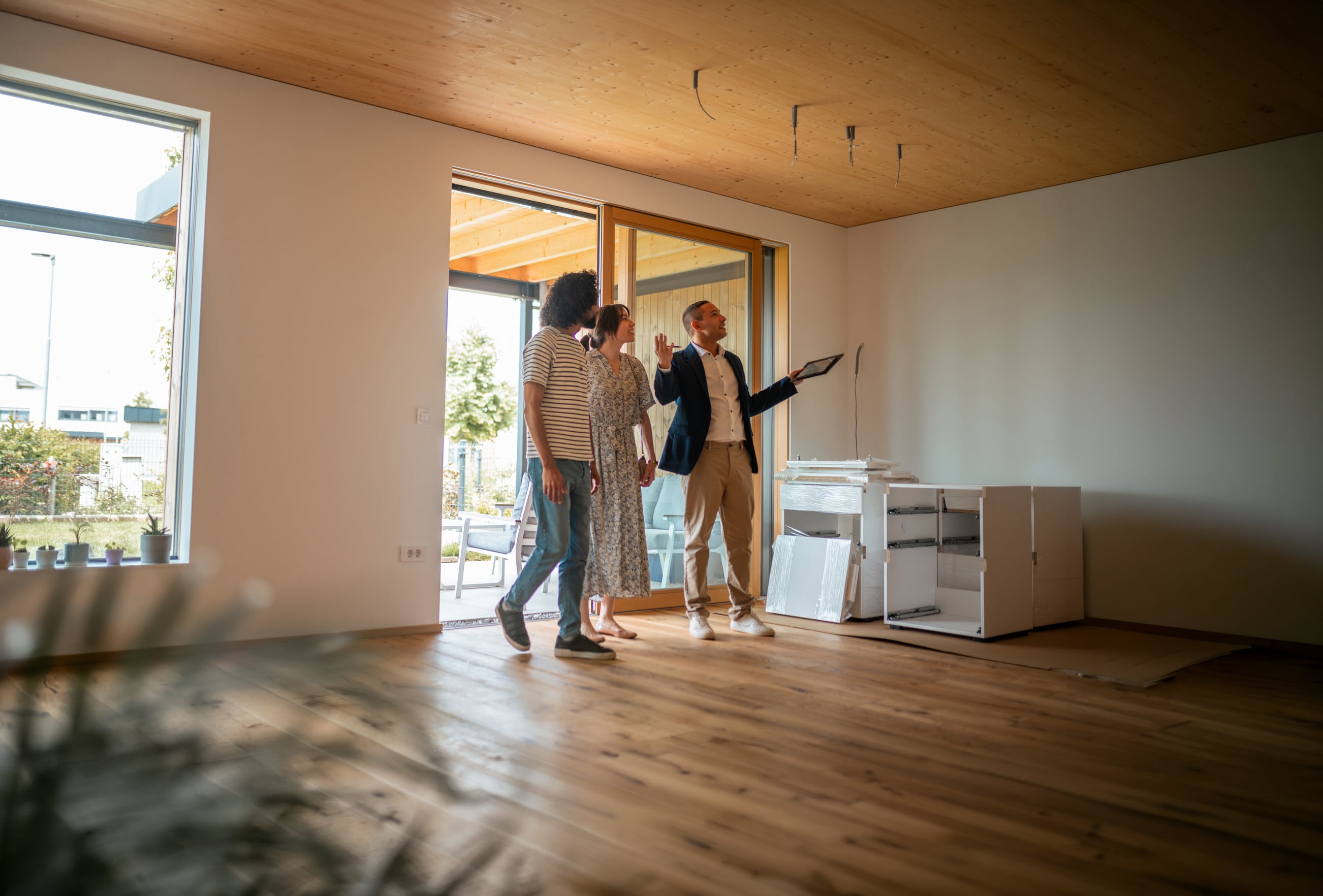 A professional Hispanic male real estate agent engages with a couple, presenting features of a spacious, sunlit home, while the diverse pair attentively follows along, considering potential ownership.