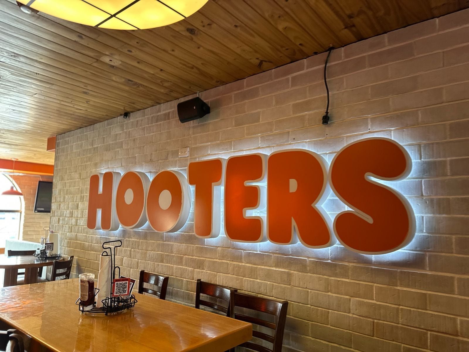 Interior de un restaurante Hooters en Costa Rica, con su característico logo iluminado en la pared de ladrillo, mesas de madera y ambiente casual.