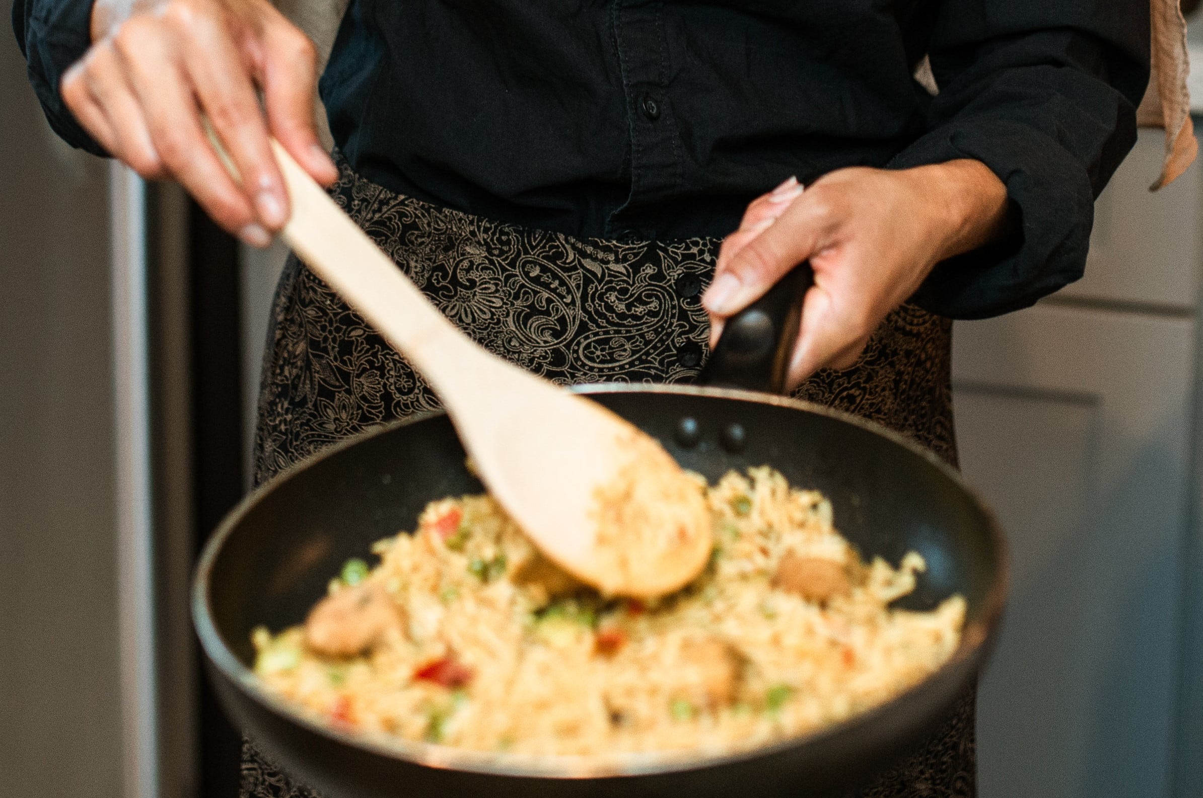 Primer plano de una persona cocinando fideos con vegetales en una sartén, usando una cuchara de madera, resaltando la preparación culinaria en contextos higiénicos.