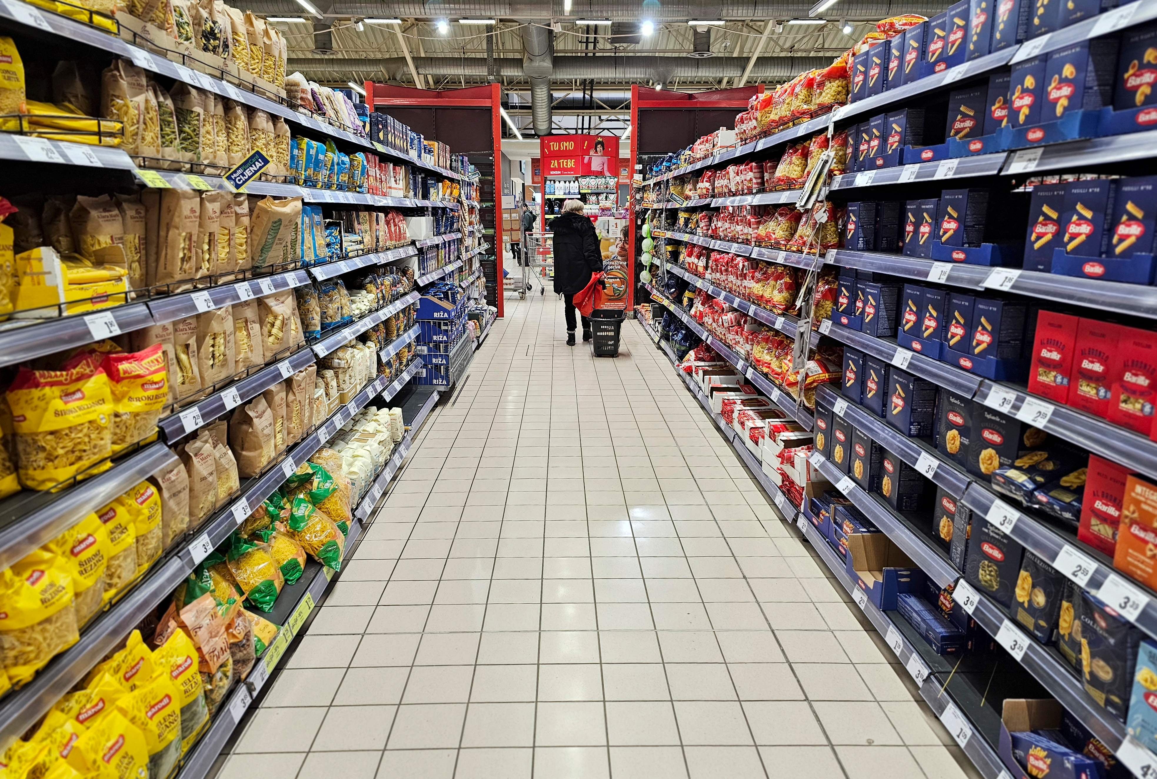 Pasillo de supermercado con estantes llenos de productos alimenticios y una persona al fondo empujando un carrito de compras.