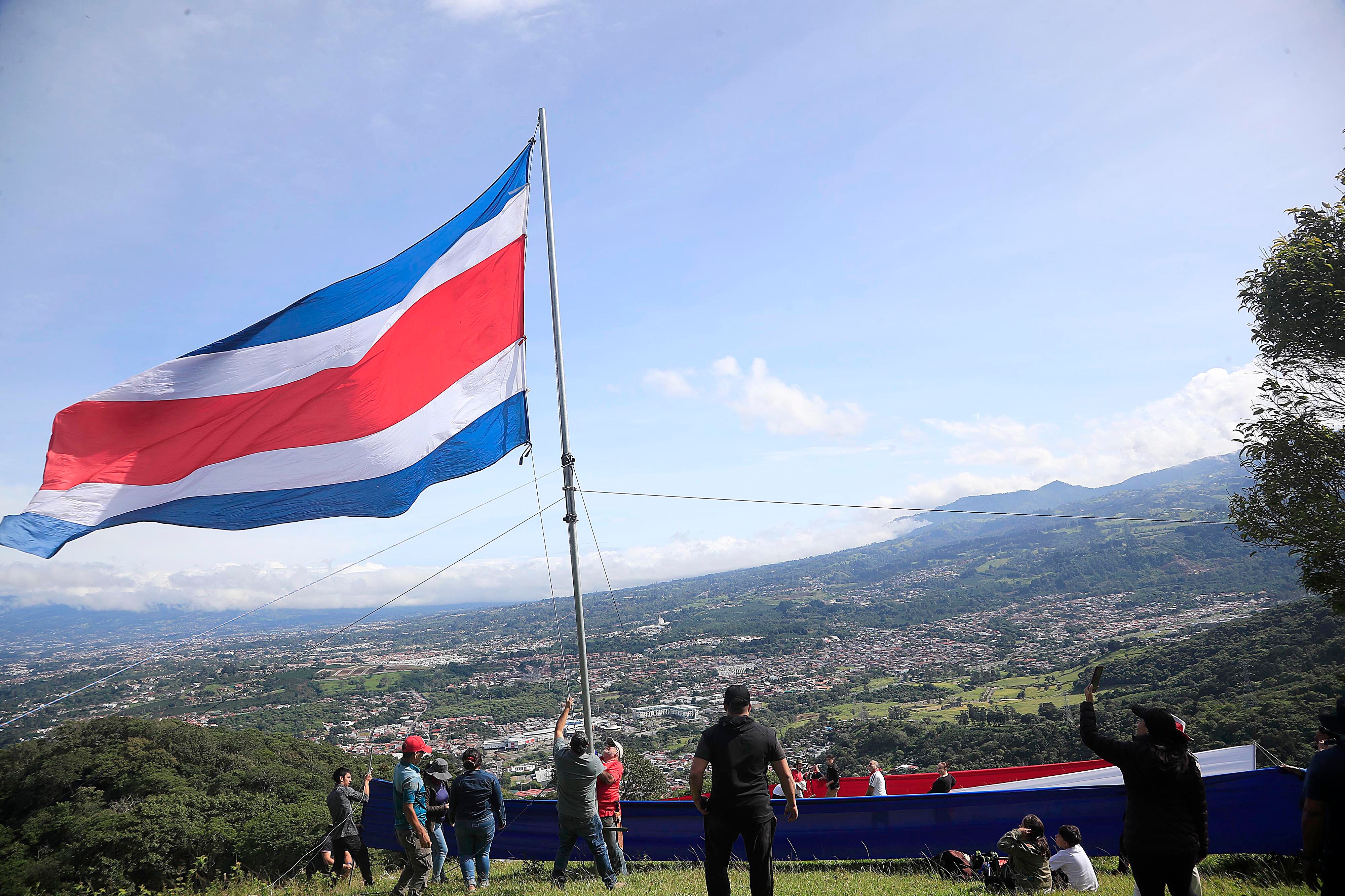 15/09/2023 Tres Ríos. Familiares y amigos de Rigoberto Vega Alvarado izaron una bandera gigante, como ha sido tradición desde hace 60 años, cada 15 de setiembre, en la ladera del potrero La Rosa, en la fila de los cerros La Carpintera. Desde las 5:00 a.m. salieron del centro del cantón de La Unión unas 30 personas, bien equipadas con herramientas, telas, tubos, cuerdas, y por supuesto la bandera. Y ya en el sitio empezaron a colocar las seis franjas de tela horizontales de 14 metros por 1 y medio de alto a una distancia de 10 metros aproximadamente cada una, de manera que desde el centro de Tres Ríos se aprecia como una sola bandera por efecto de la aproximación de los planos. Todos trabajan porque mientras unos se ocupan de colocar las franjas individuales de tela, otros trabajan en la colocación del tubo gigante para luego izar la bandera, y otros más, especialmente las mujeres se ocupan de hacer el café, la aguadulce y los sandwiches. Mientras se trabaja en el levantamiento de la bandera y la colocación de las telas individuales las solo el viento compite con las notas de canciones tradicionales del folklor costarricense propias de la época que salen de un enorme parlante.