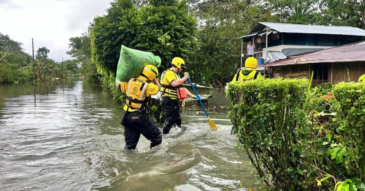 Dos rescatistas con uniformes amarillos cruzan una calle inundada llevando suministros de ayuda, frente a una vivienda afectada por las lluvias en Costa Rica.