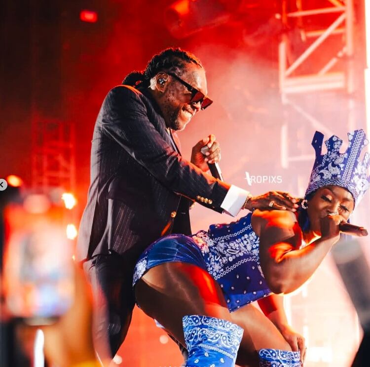 Vybz Kartel cantando en concierto, con el micrófono en mano.  usa un traje de color negro y corbata dorada.