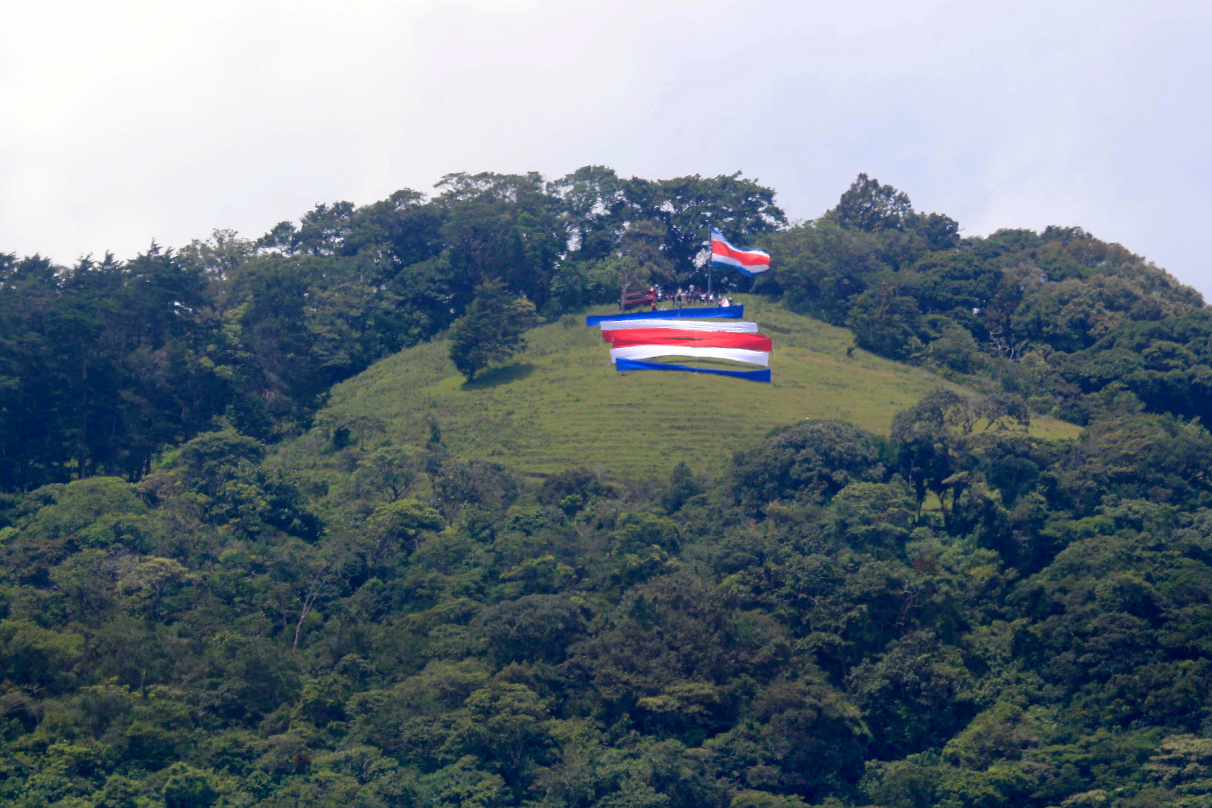 15/09/2023 Tres Ríos. Familiares y amigos de Rigoberto Vega Alvarado izaron una bandera gigante, como ha sido tradición desde hace 60 años, cada 15 de setiembre, en la ladera del potrero La Rosa, en la fila de los cerros La Carpintera. Desde las 5:00 a.m. salieron del centro del cantón de La Unión unas 30 personas, bien equipadas con herramientas, telas, tubos, cuerdas, y por supuesto la bandera. Y ya en el sitio empezaron a colocar las seis franjas de tela horizontales de 14 metros por 1 y medio de alto a una distancia de 10 metros aproximadamente cada una, de manera que desde el centro de Tres Ríos se aprecia como una sola bandera por efecto de la aproximación de los planos. Todos trabajan porque mientras unos se ocupan de colocar las franjas individuales de tela, otros trabajan en la colocación del tubo gigante para luego izar la bandera, y otros más, especialmente las mujeres se ocupan de hacer el café, la aguadulce y los sandwiches. Mientras se trabaja en el levantamiento de la bandera y la colocación de las telas individuales las solo el viento compite con las notas de canciones tradicionales del folklor costarricense propias de la época que salen de un enorme parlante.