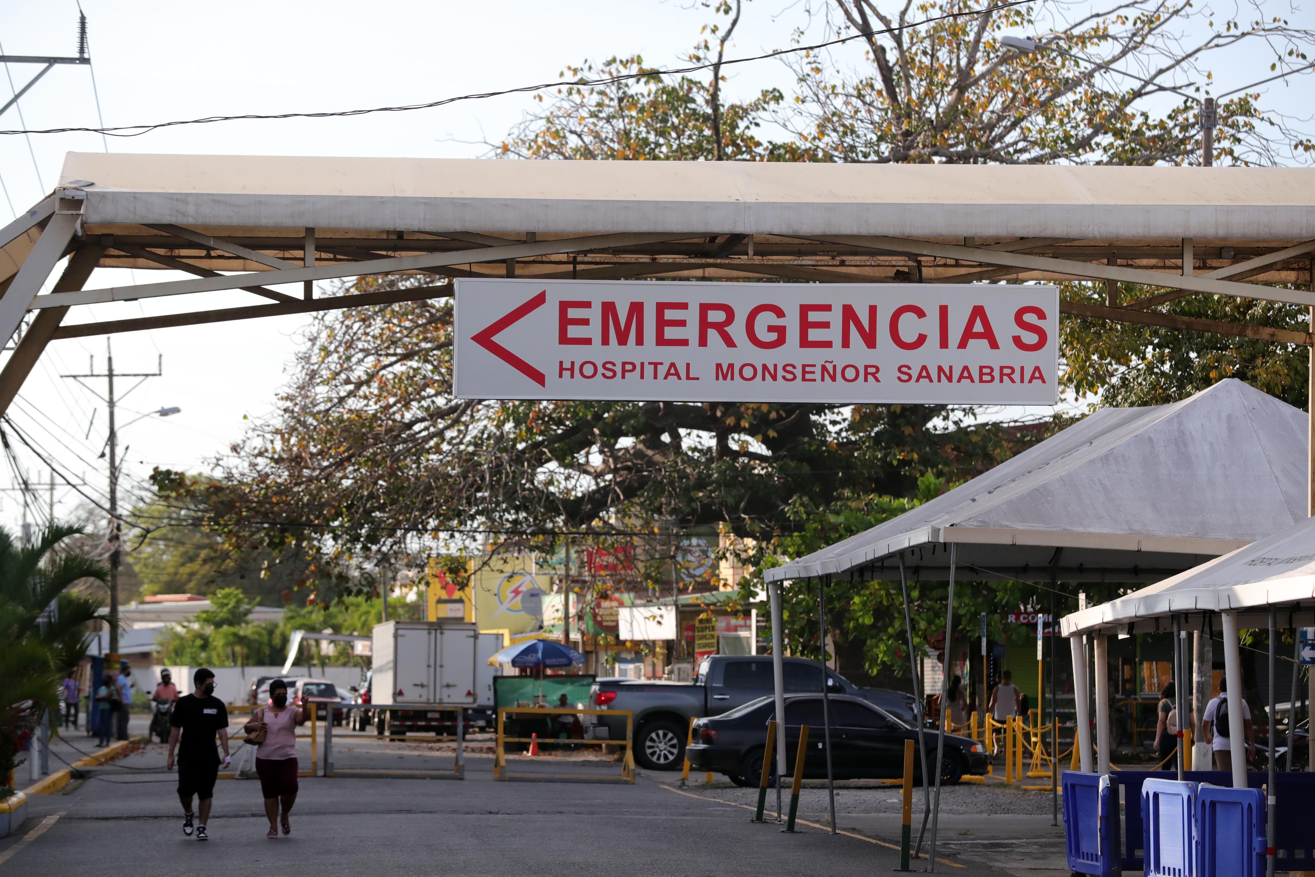 Letrero de la entrada del área de emergencias del Hospital Monseñor Sanabria en Puntarenas, con personas caminando cerca y carpas en el entorno.