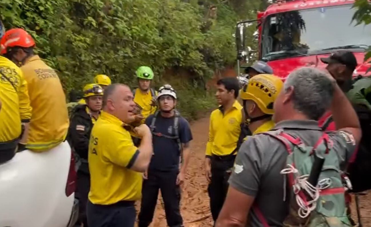 Socorristas de la Cruz Roja y bomberos trabajando en el sitio del accidente de una avioneta en los cerros de Escazú, Costa Rica. La escena incluye personal de rescate con cascos y equipo especializado junto a un camión de bomberos.