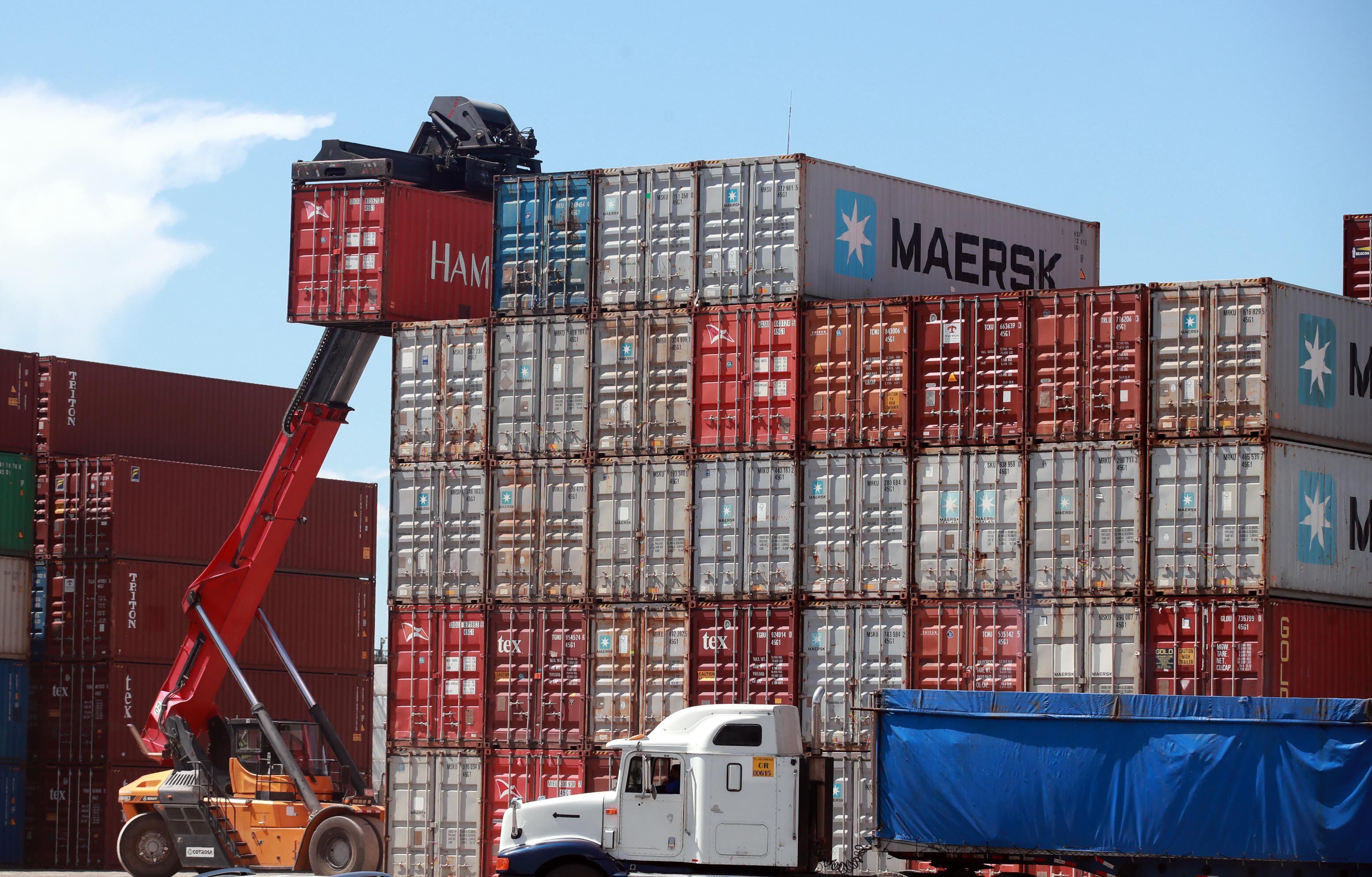Montacargas trasladando un contenedor rojo en un puerto comercial con pilas de contenedores de diferentes colores al fondo. Un camión de carga espera en primer plano.